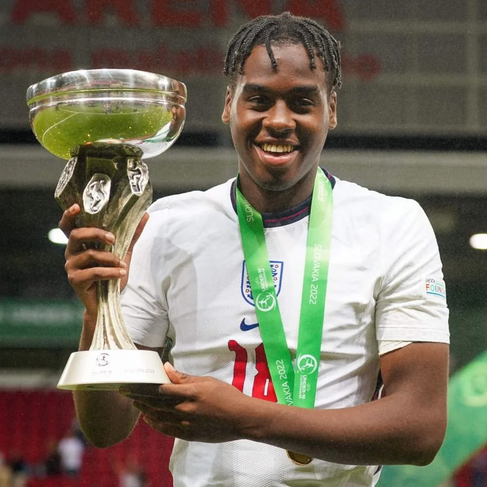 A young man celebrating and smiling while holding a trophy after a sports event, wearing a white sports jersey and a green winner's medal around his neck.