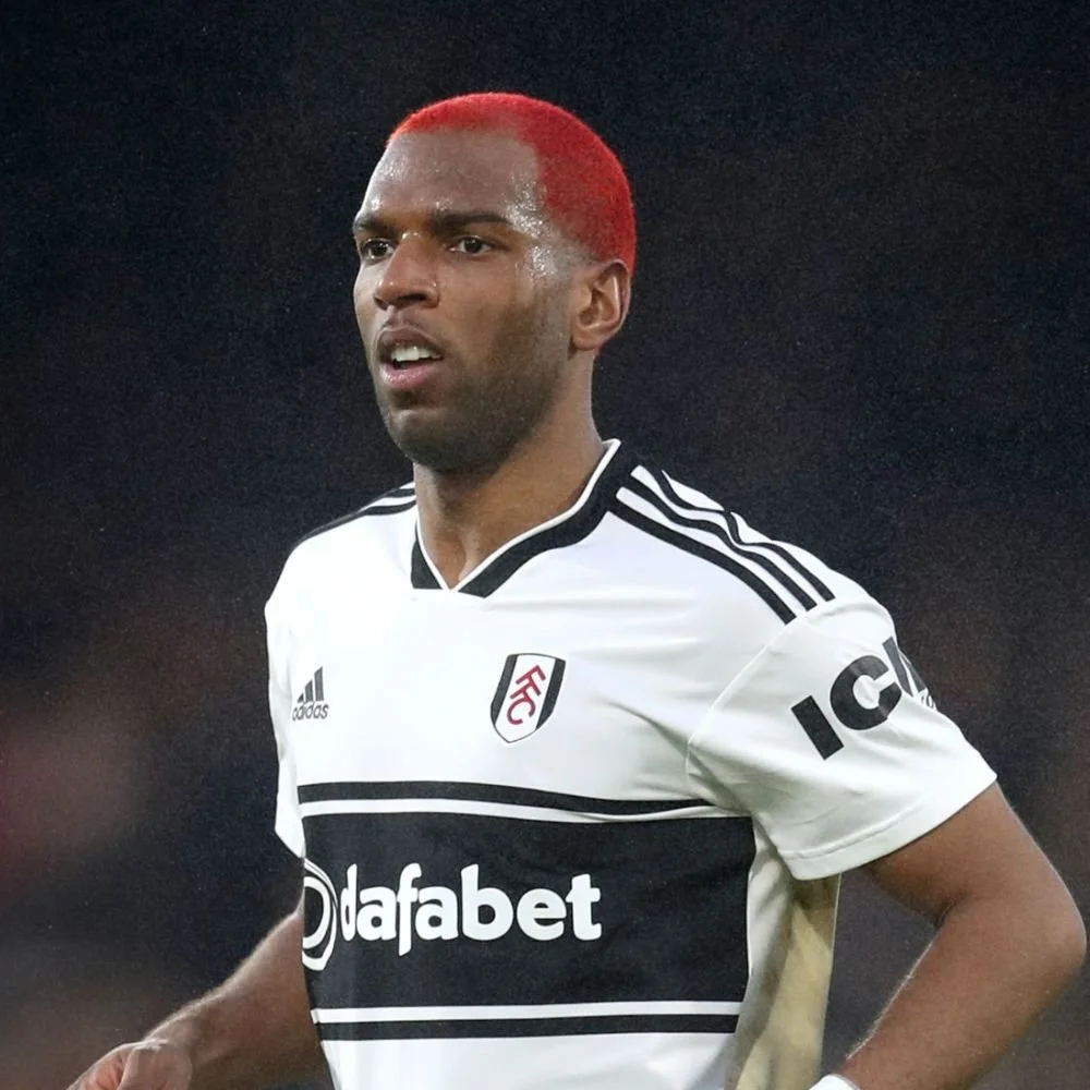 A football player with bright red hair wearing a white and black jersey with sponsor logos, on a dark background.