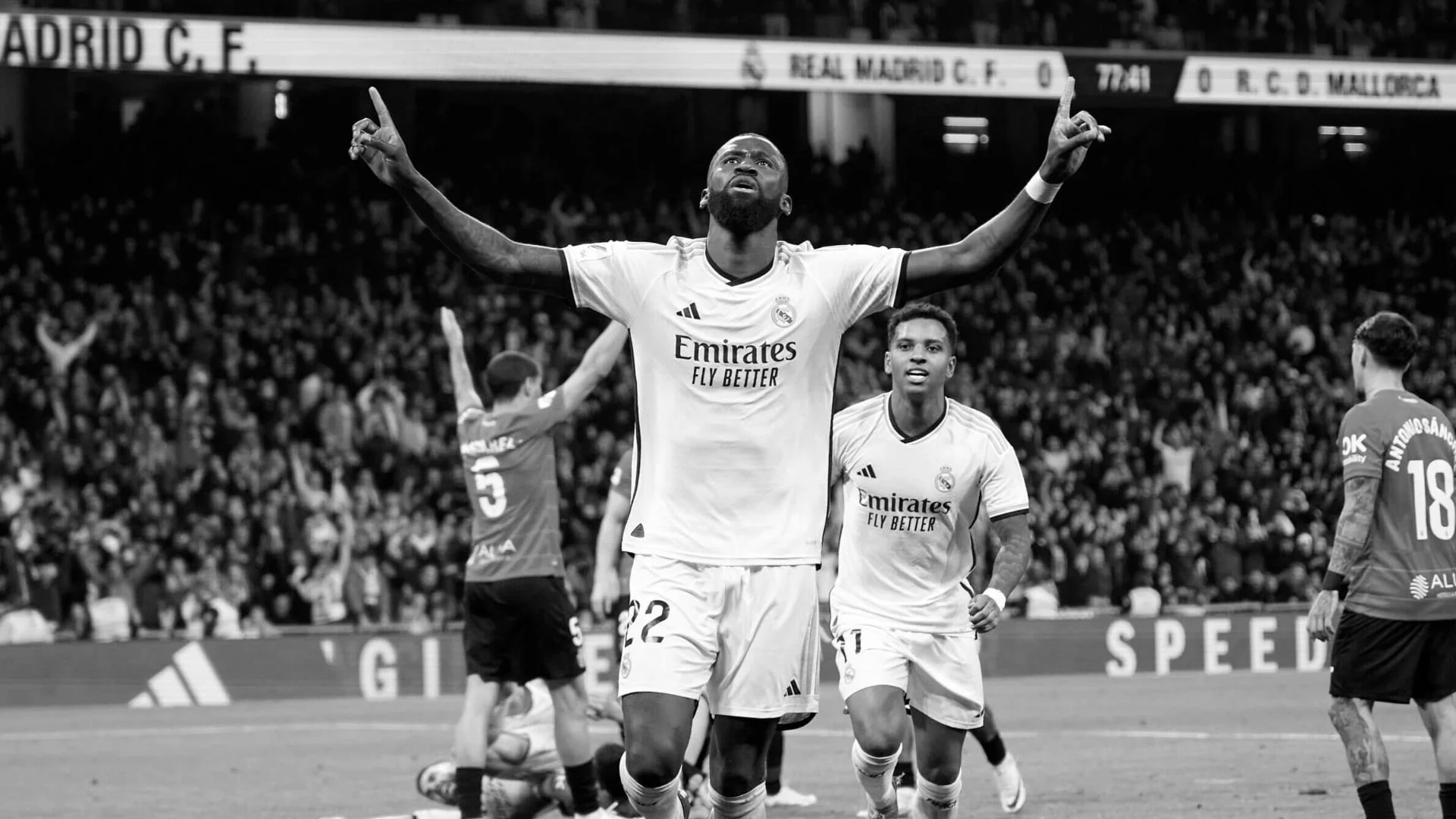 Black and white photo of a soccer player in a white Real Madrid jersey celebrating with arms raised, teammates around him, crowds in the background.