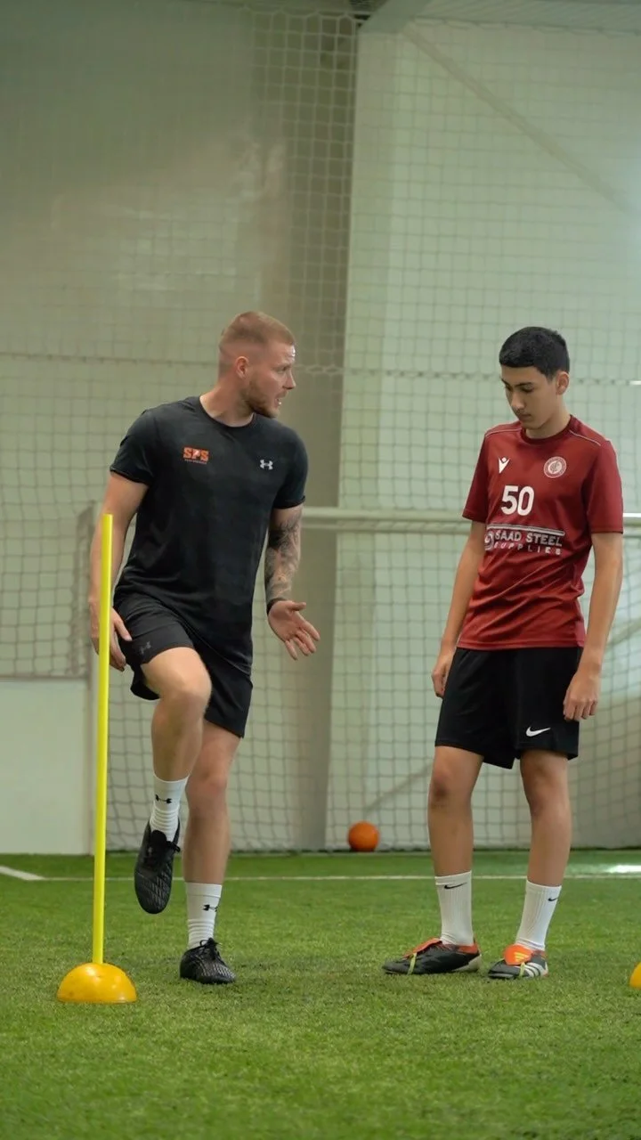 A coach and a young soccer player are in an indoor training facility. The coach, wearing black sportswear, appears to be instructing or explaining something to the player, who is dressed in a red jersey and black shorts. The player is standing with a serious expression, and there are soccer training poles and cones on the turf.