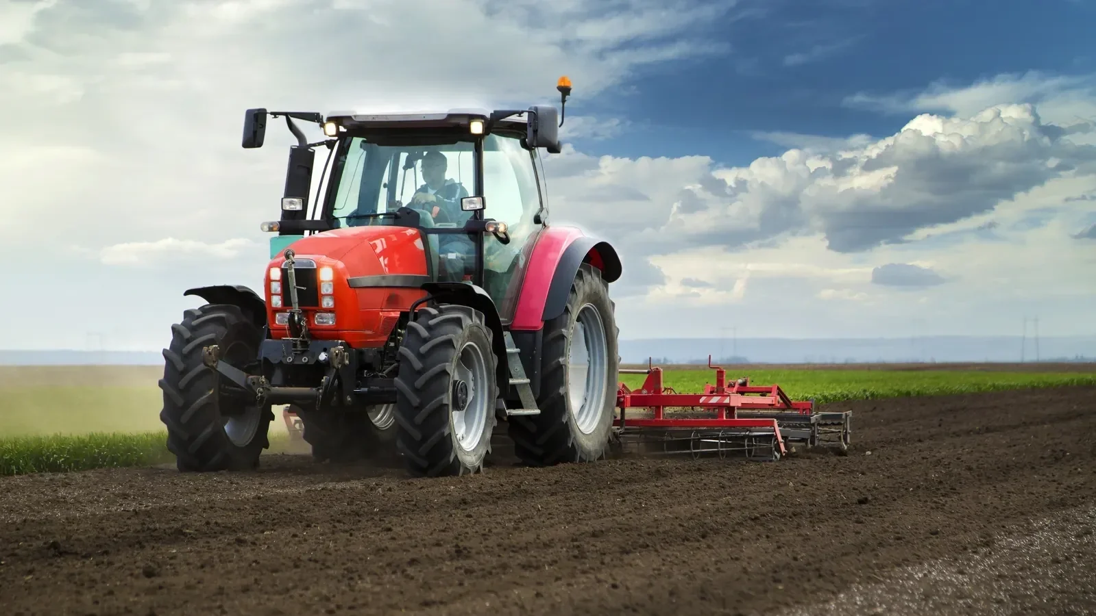 Red tractor with a farm worker onboard, tilling dark brown soil on a vast farmland under a cloudy sky.