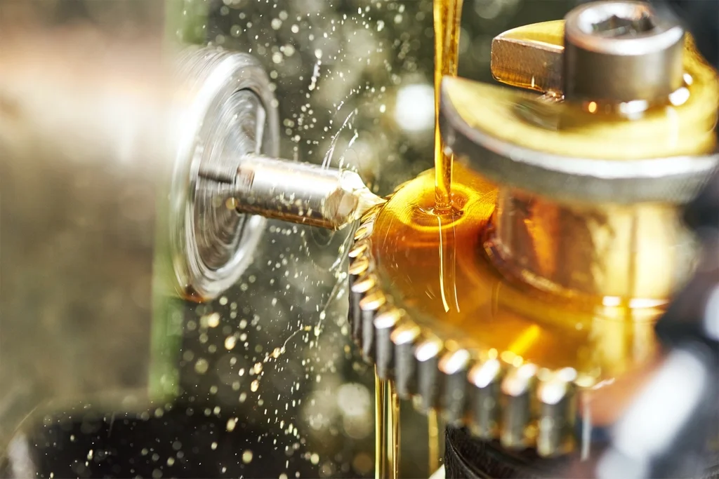 Close-up of a metal gear being lubricated with golden oil, with oil pouring from a spout.