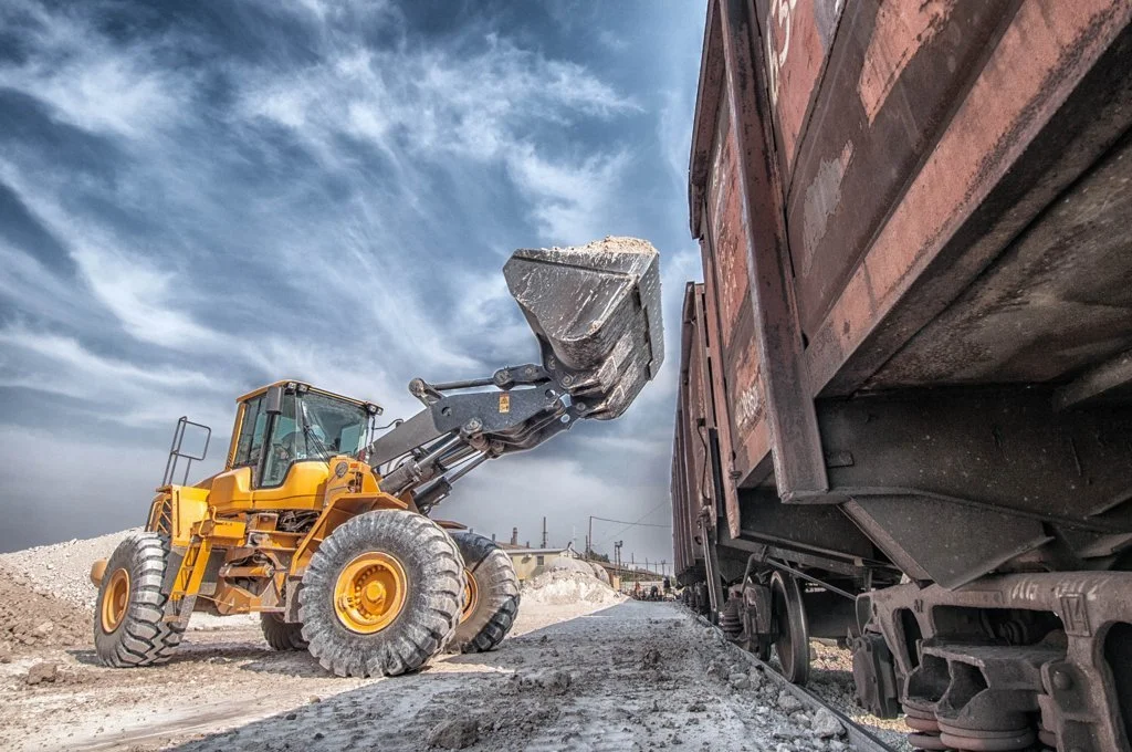 A large yellow front loader is scooping gravel or dirt near a train car on a construction or industrial site under a partly cloudy sky.