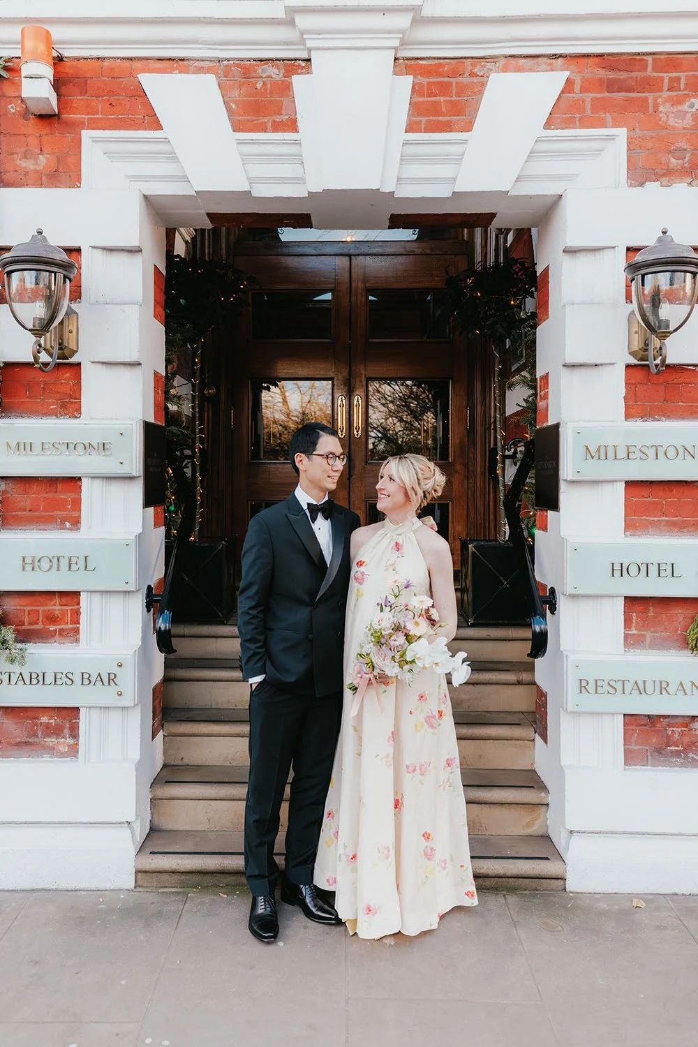 ウエディングドレスを着た女性とタキシードを着た男性がお互いに見つめ合っている。背景は赤レンガの建物と木製のドア。