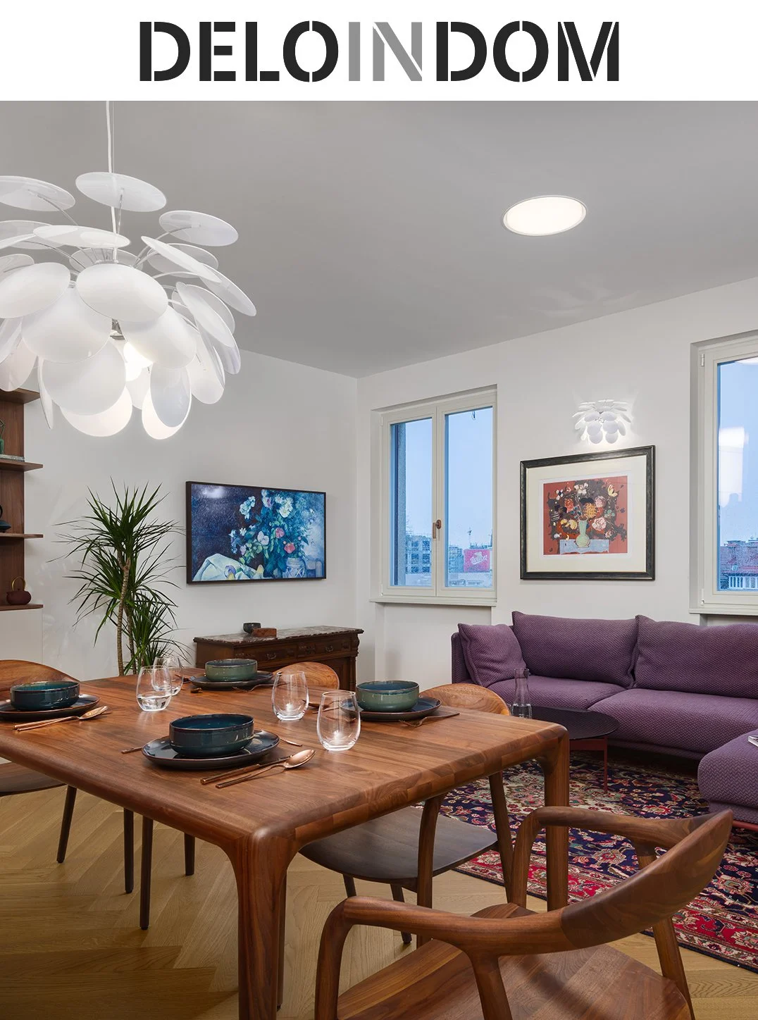 A modern dining and living room with a wooden dining table set with bowls, glasses, and cutlery, a purple sofa, framed artwork on white walls, a potted plant, and large windows showing city buildings.