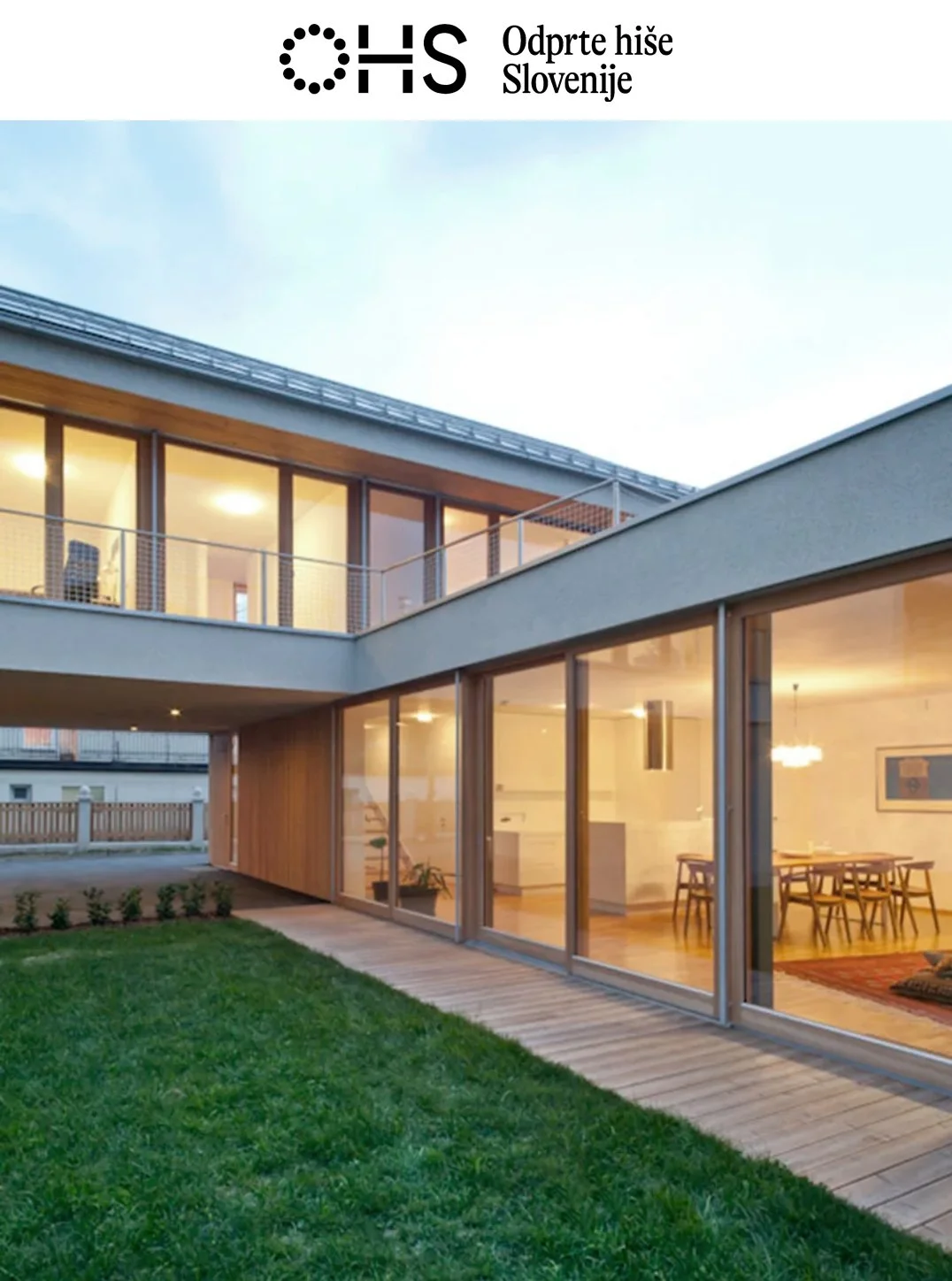 A modern two-story house with large glass windows and wooden accents, featuring an interior dining area visible through the glass, a green lawn, and a wooden pathway outside, during dusk.