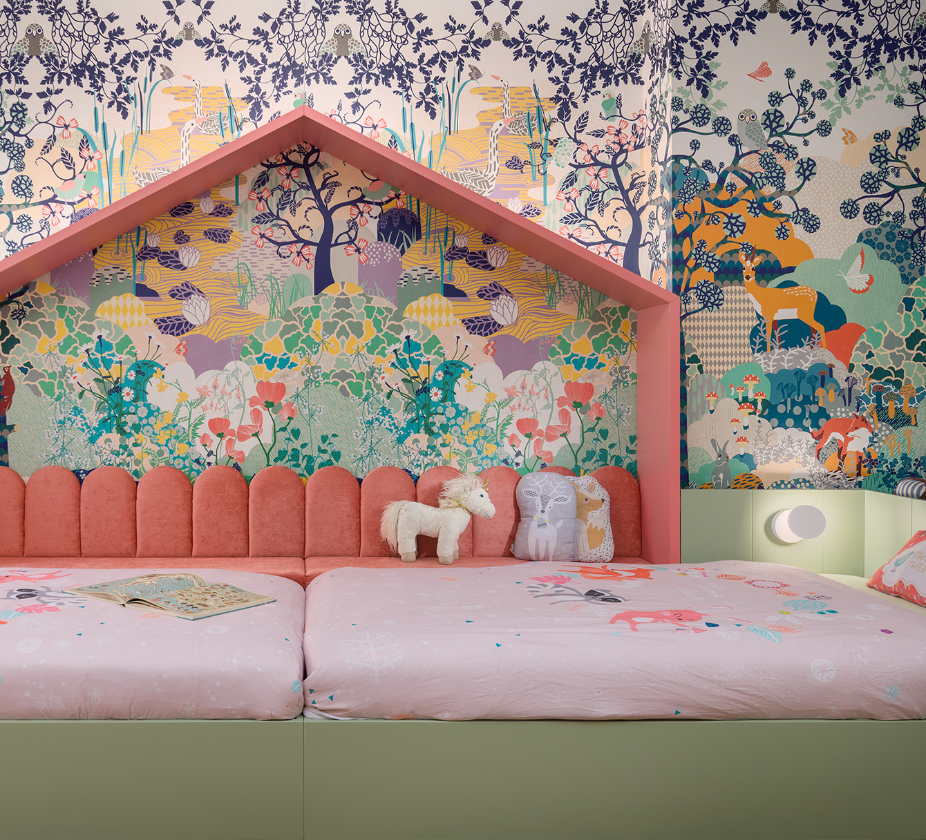 A bed with a pink upholstered headboard and wallpaper featuring a colorful jungle scene with animals and trees. There are plush toys, including a white horse and owl pillows, and an open book on the bed. Next to the bed is a light fixture with a white and pink spherical shade.
