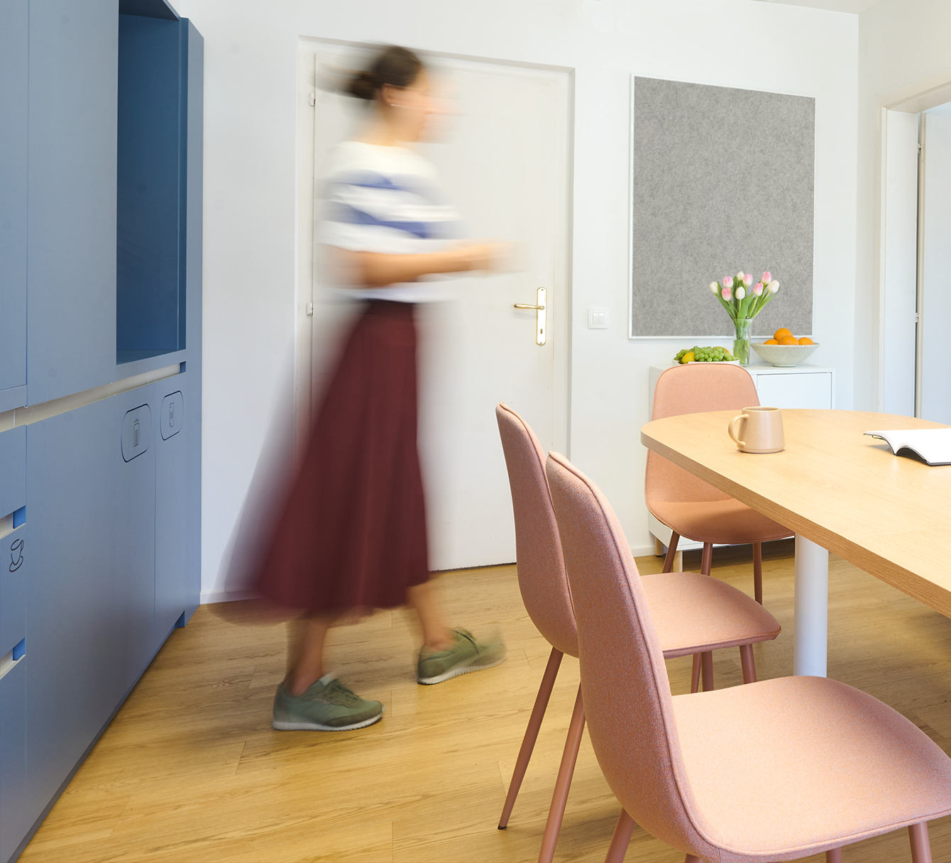 A woman walking in a bright, modern room with pink chairs, a wooden table, a white cabinet with a vase of pink tulips, and bowls of fruit. The woman is blur due to motion.