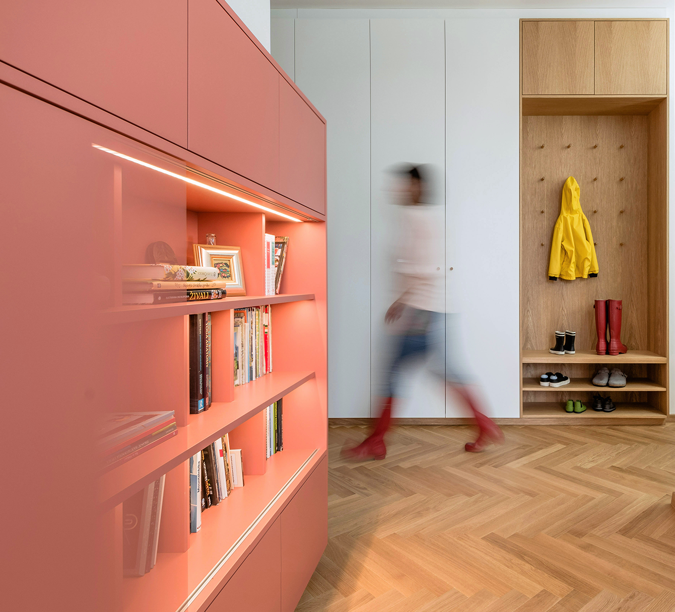 A blurred child walking through a contemporary room with a pink bookshelf filled with books on the left and a wooden coat rack with children's clothing and shoes on the right.