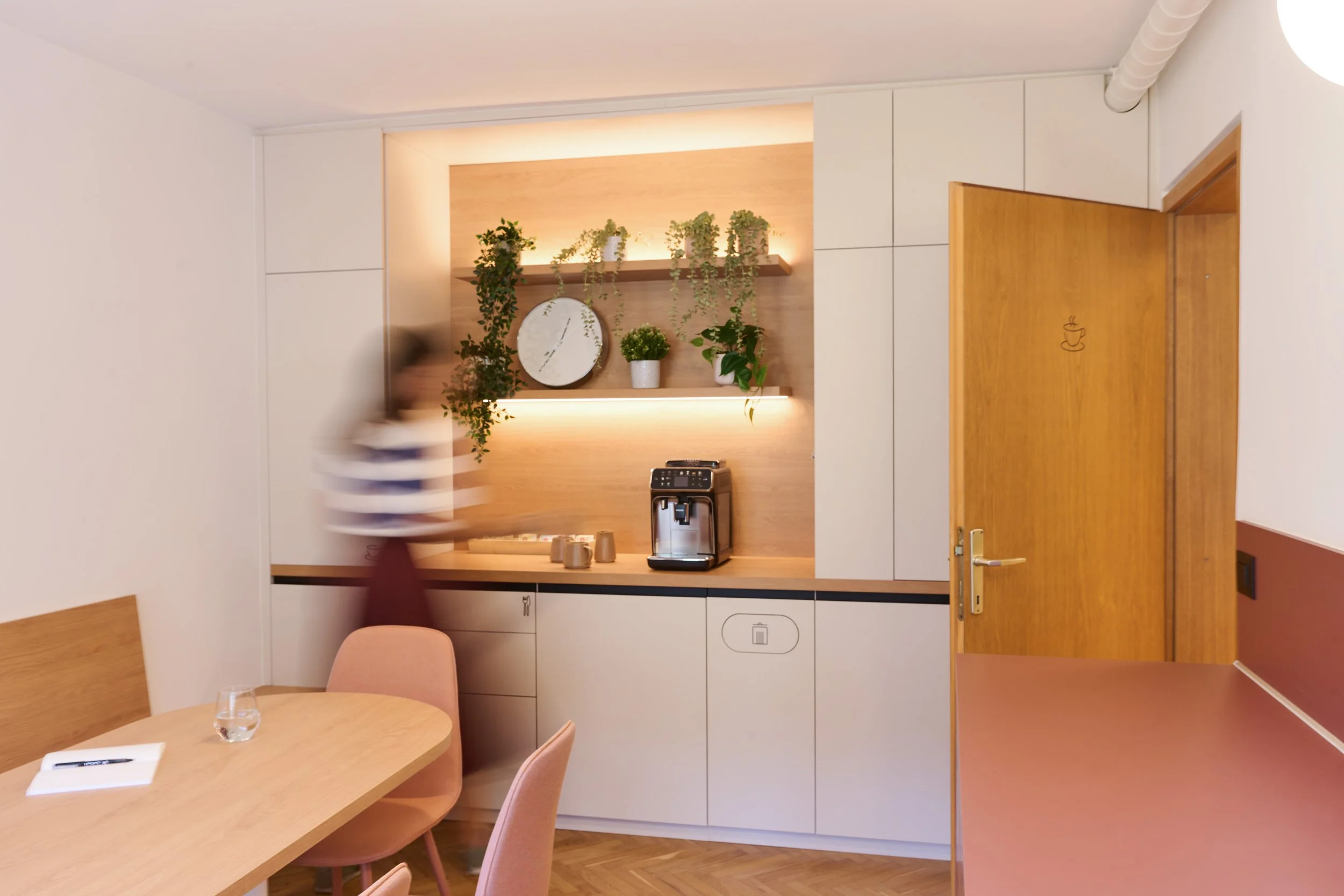 Kitchen corner with a wooden countertop, plants on wall shelf, coffee machine, cups, and a closed wooden door.