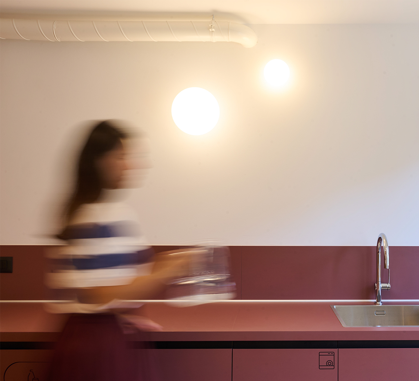 A blurry woman with dark hair, wearing a striped top, holds a plate and glass in a modern kitchen with a maroon lower wall and a stainless steel sink, illuminated by two bright white circular lights on a white wall.