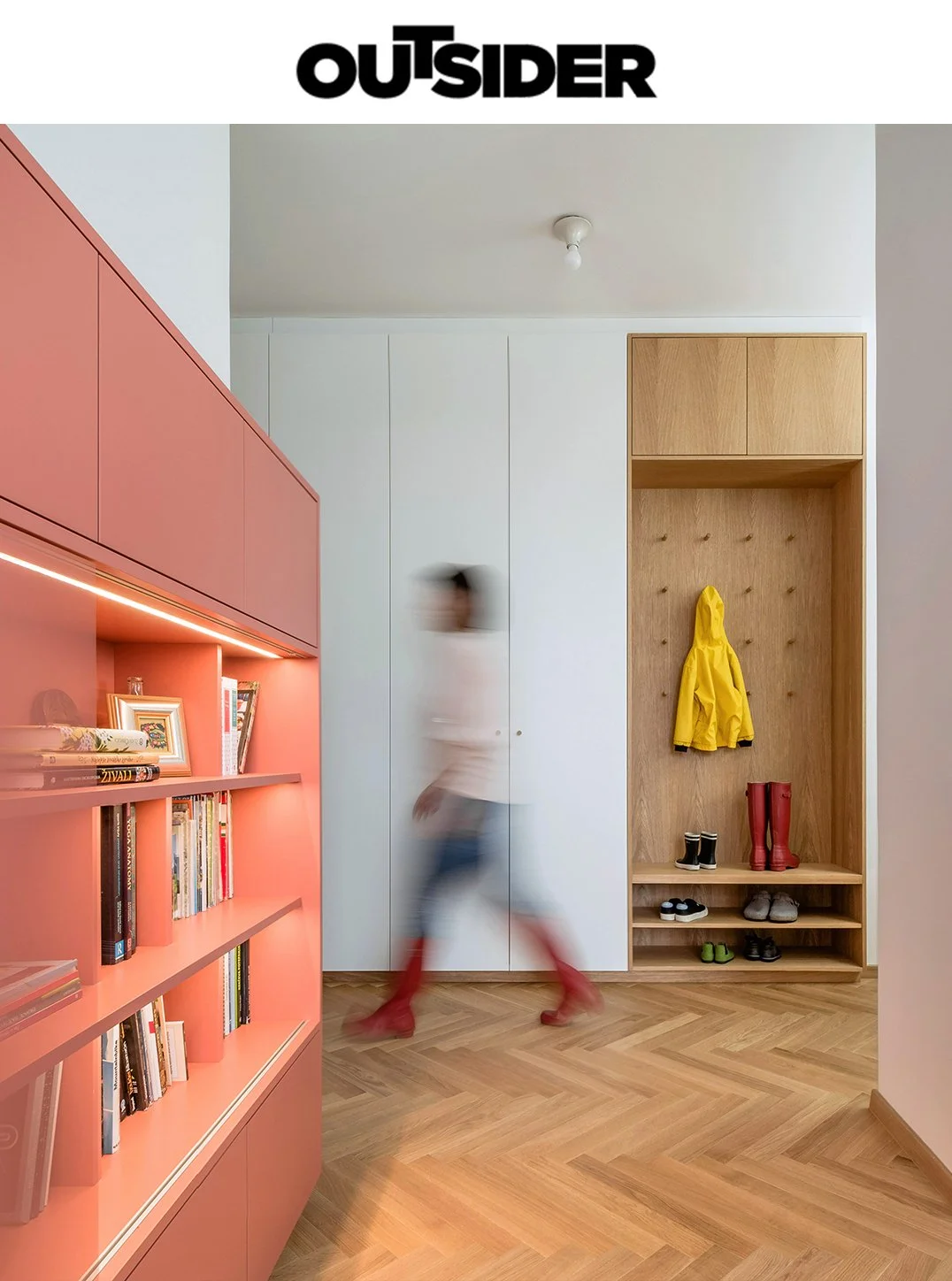 A blurred person walking in a modern, colorful hallway with wooden floor and built-in storage spaces, including a pink bookshelf on the left and a wooden coat rack with shoes on the right.