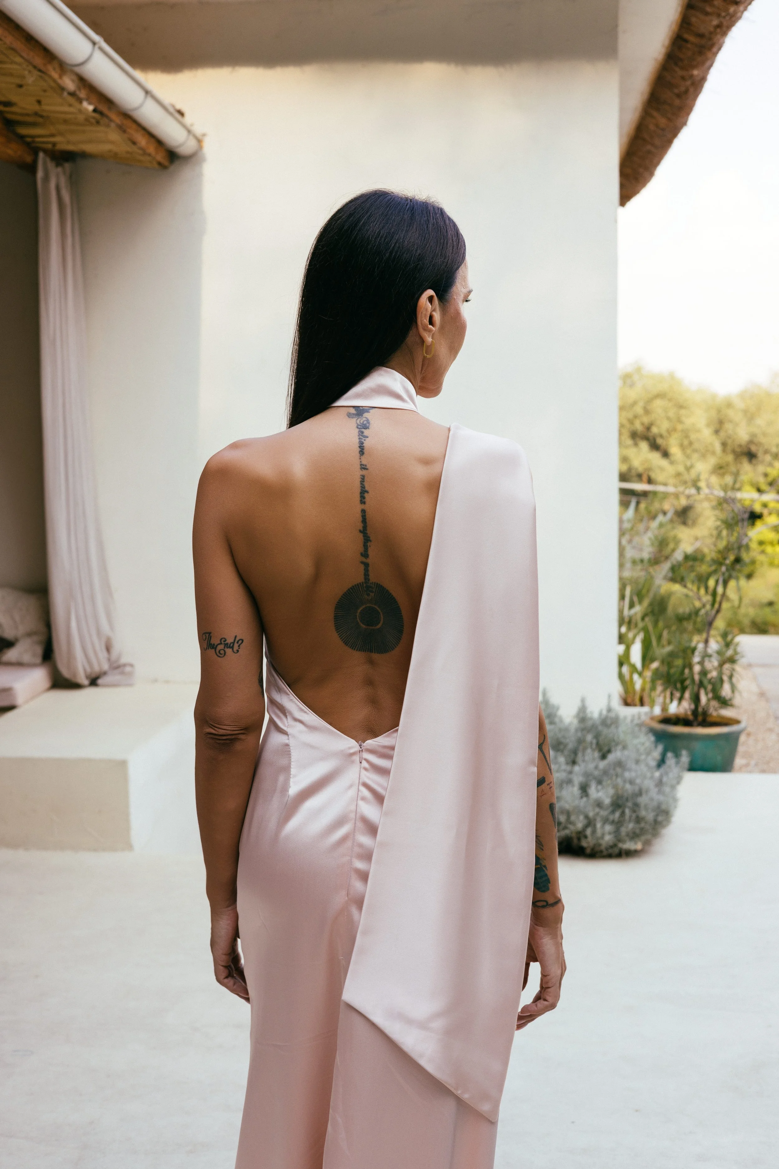 Žena u svilenkastoj, bledo roze haljini bez leđa, sa dugom crnom kosom i tetovažama na ruci i leđima, stoji na terasi i gleda ka bašti punoj drveća i biljaka. Bobana Simovic