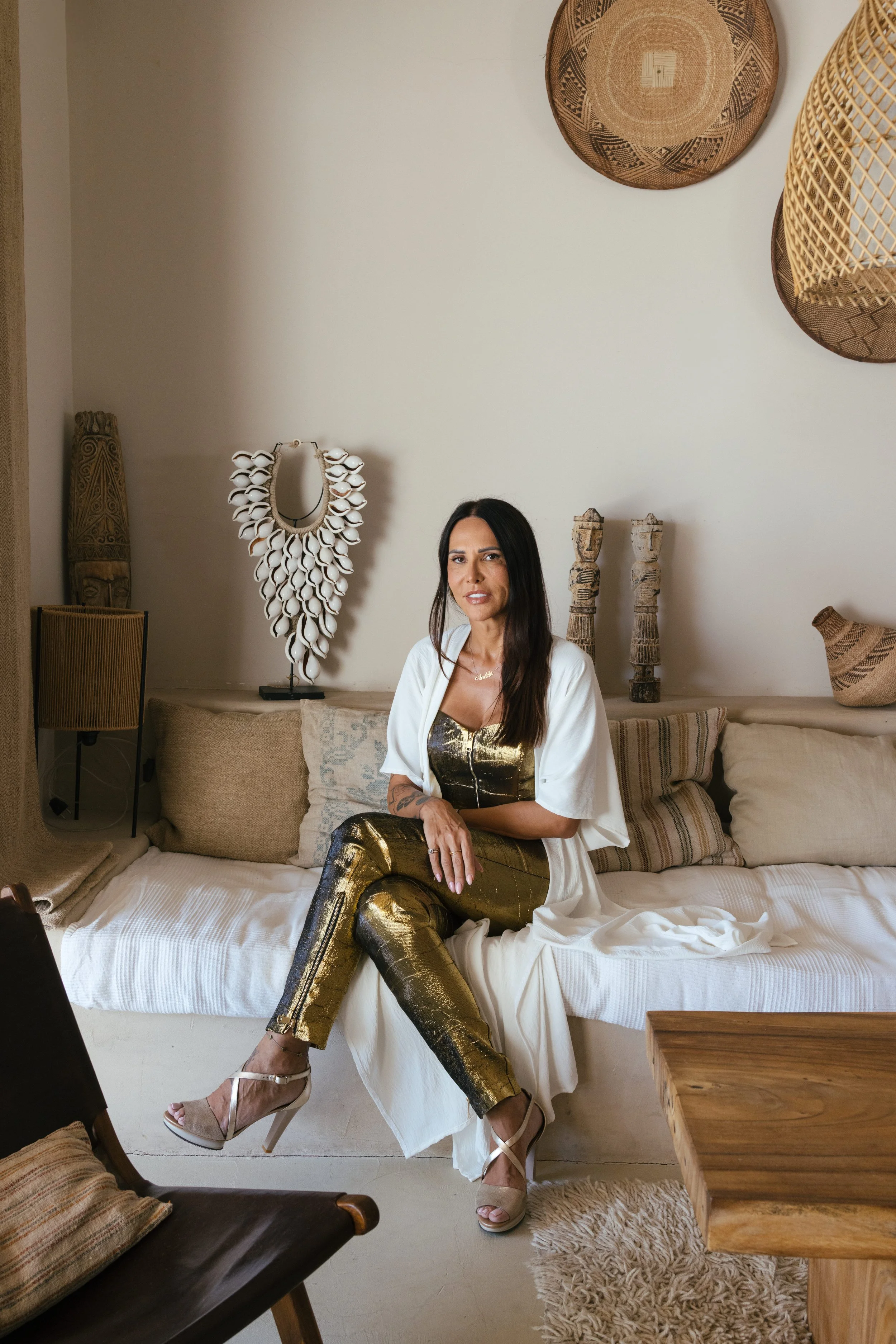 Žena sedi na bež sofi u prostoriji boho stila sa pletenim korpama i tribal umetnošću, obučena u zlatno-crne pantalone sa šarom, zlatni top i bež štikle, obasjana prirodnim svetlom. Bobana Simovic