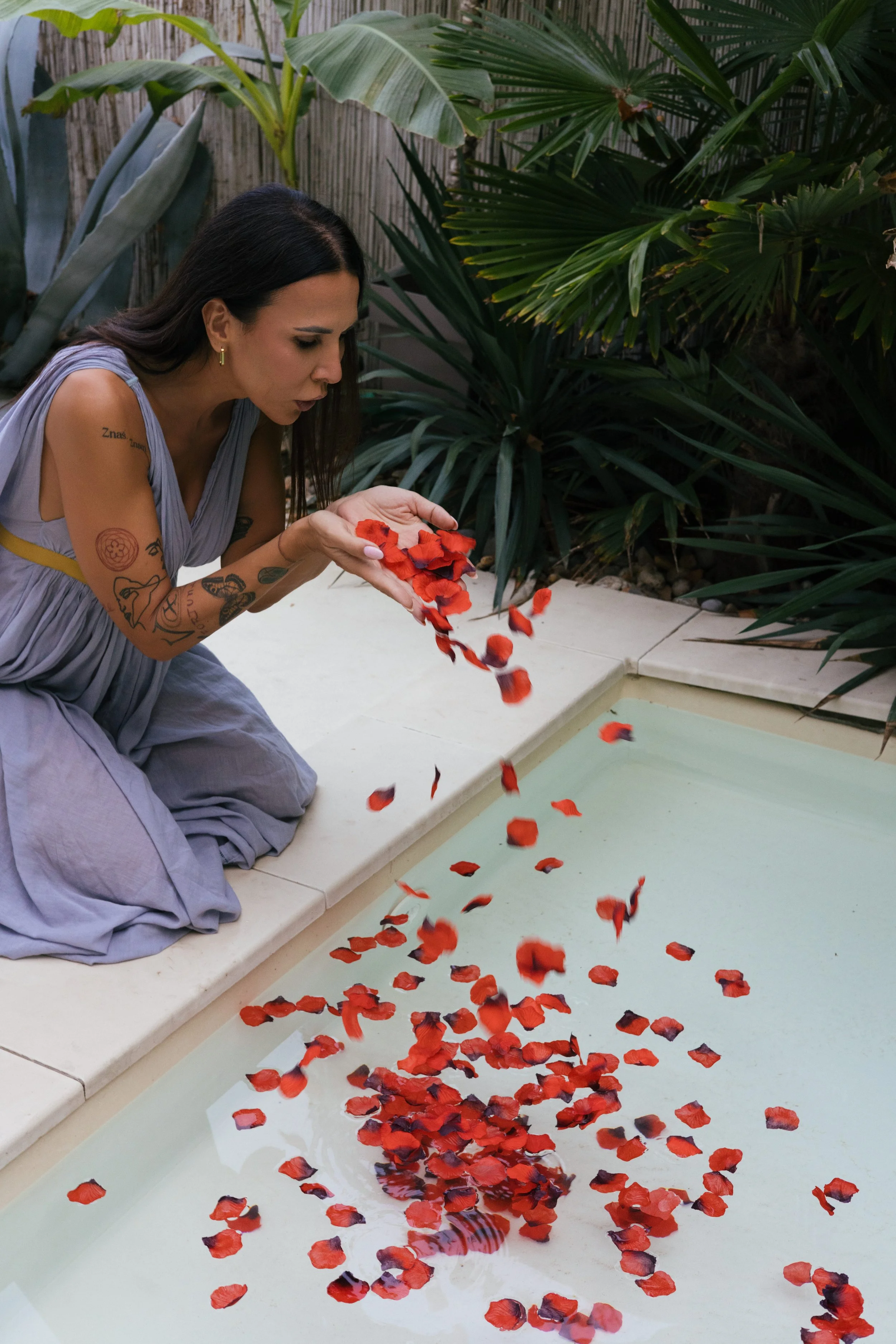 Žena u sivoj haljini, sa tetovažama na ruci, kleči pored bazena i posipa crvene latice ruža u vodu, okružena bujnim zelenim tropskim biljkama. Bobana Simovic