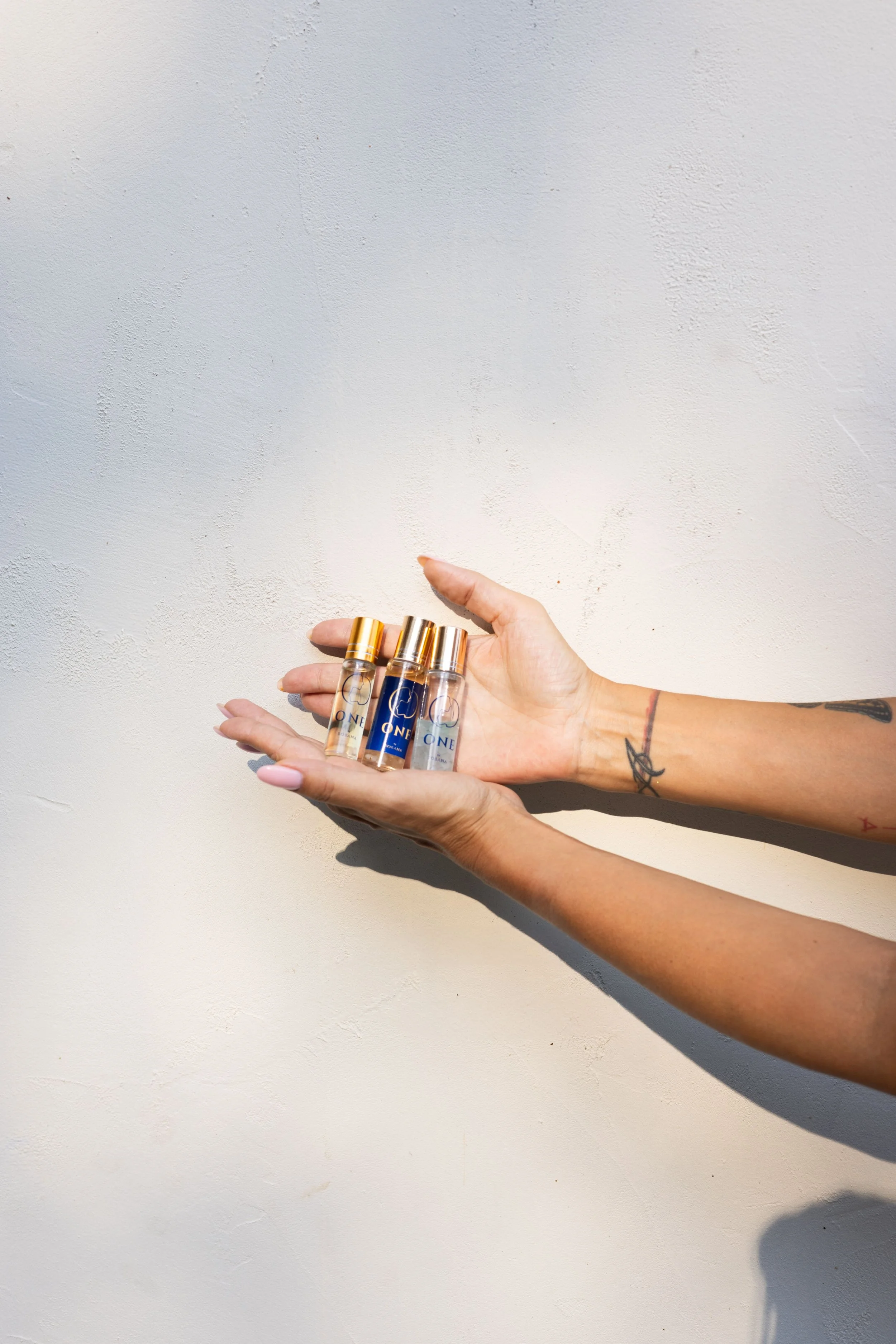 Ruka sa tetovažama drži tri male bočice sa zlatnim čepovima i plavim etiketama, naspram bele podloge — trenutak čistote, istine i dodira prirodne esencije.
