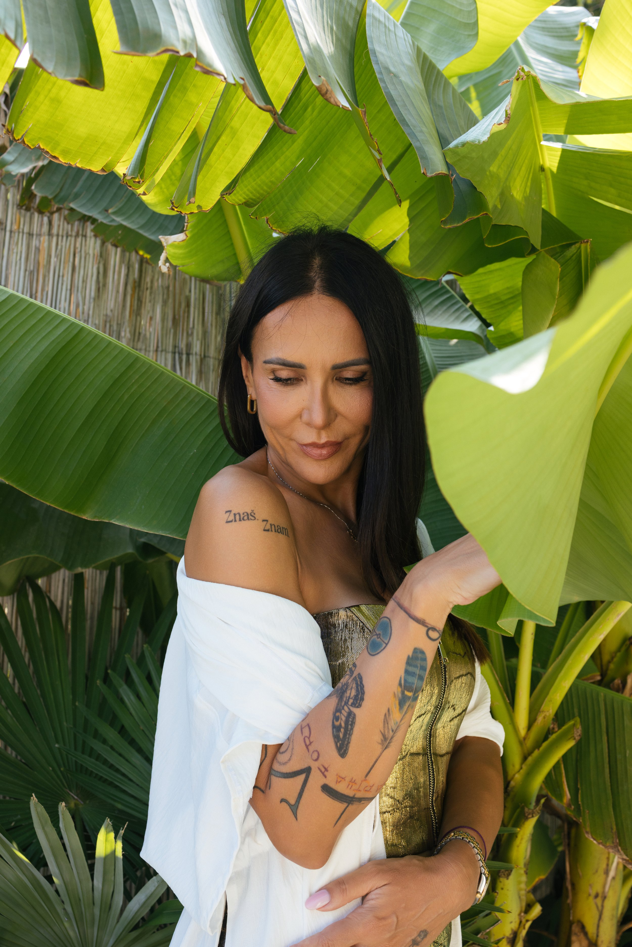 Žena tamne kose i tetovaža na ruci, u beloj bluzi spuštenih ramena, stoji među velikim zelenim listovima banane, opuštenog i zamišljenog izraza. Bobana Simovic