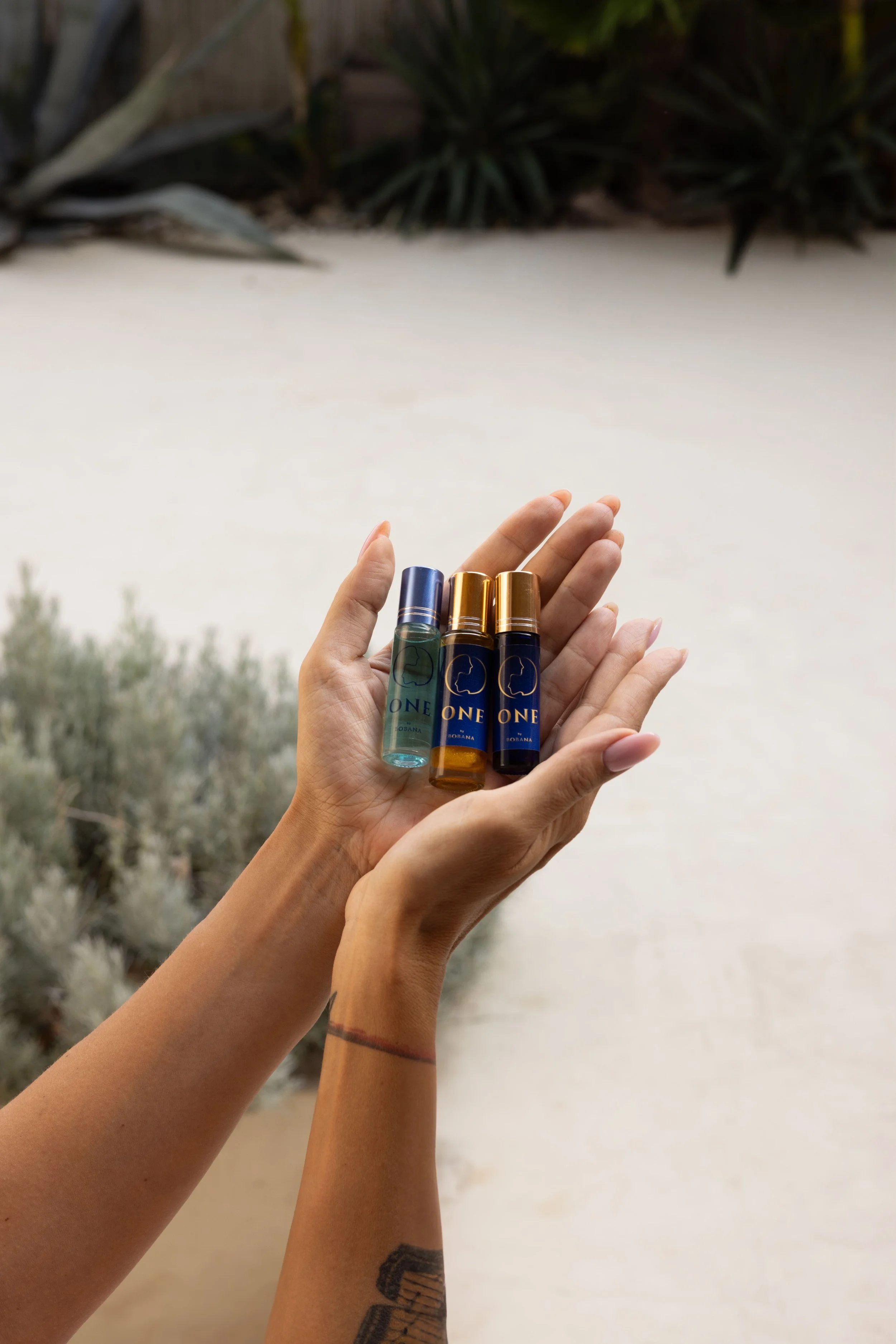 Ruka koja drži tri parfemske bočice sa plavim i zlatnim čepovima, naspram neutralne pozadine sa nežnim zelenilom u pozadini — spoj elegancije, prirode i mirisne istine.