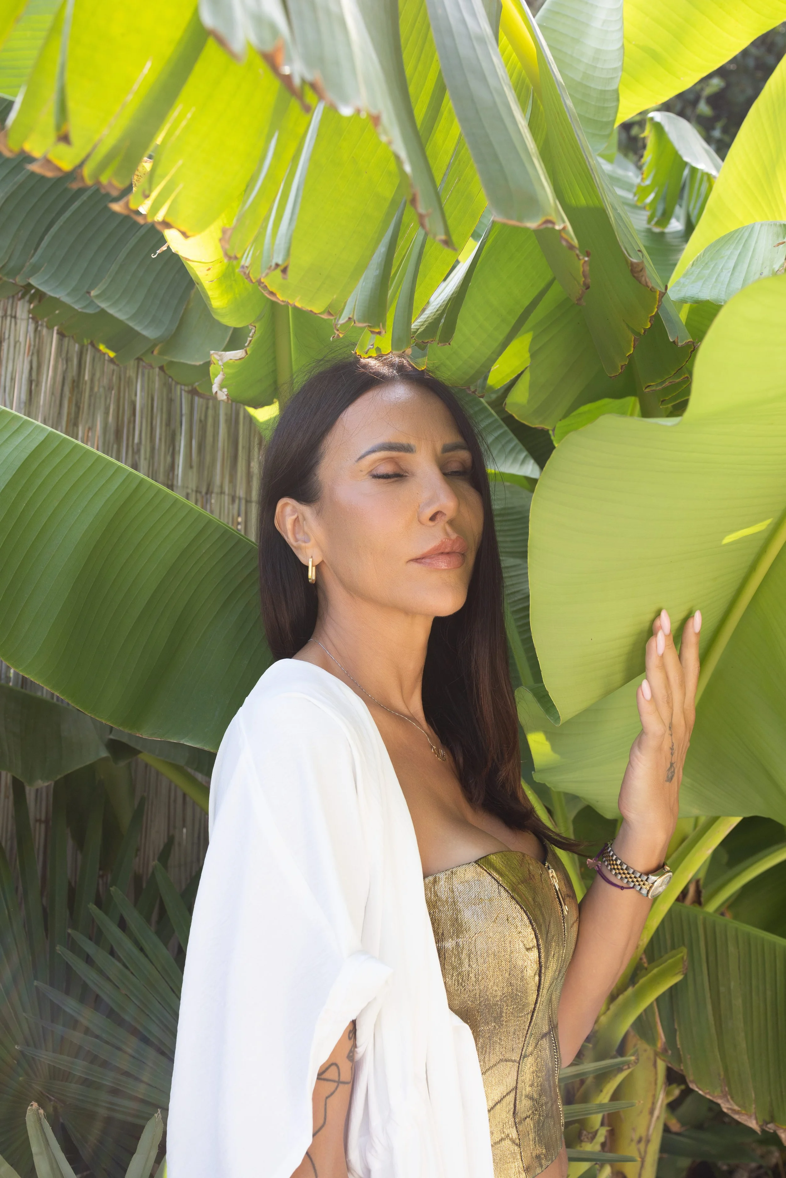 Žena tamne kose, sa nakitom, koja naslanja ruku na veliki zeleni list banane među tropskim biljkama.