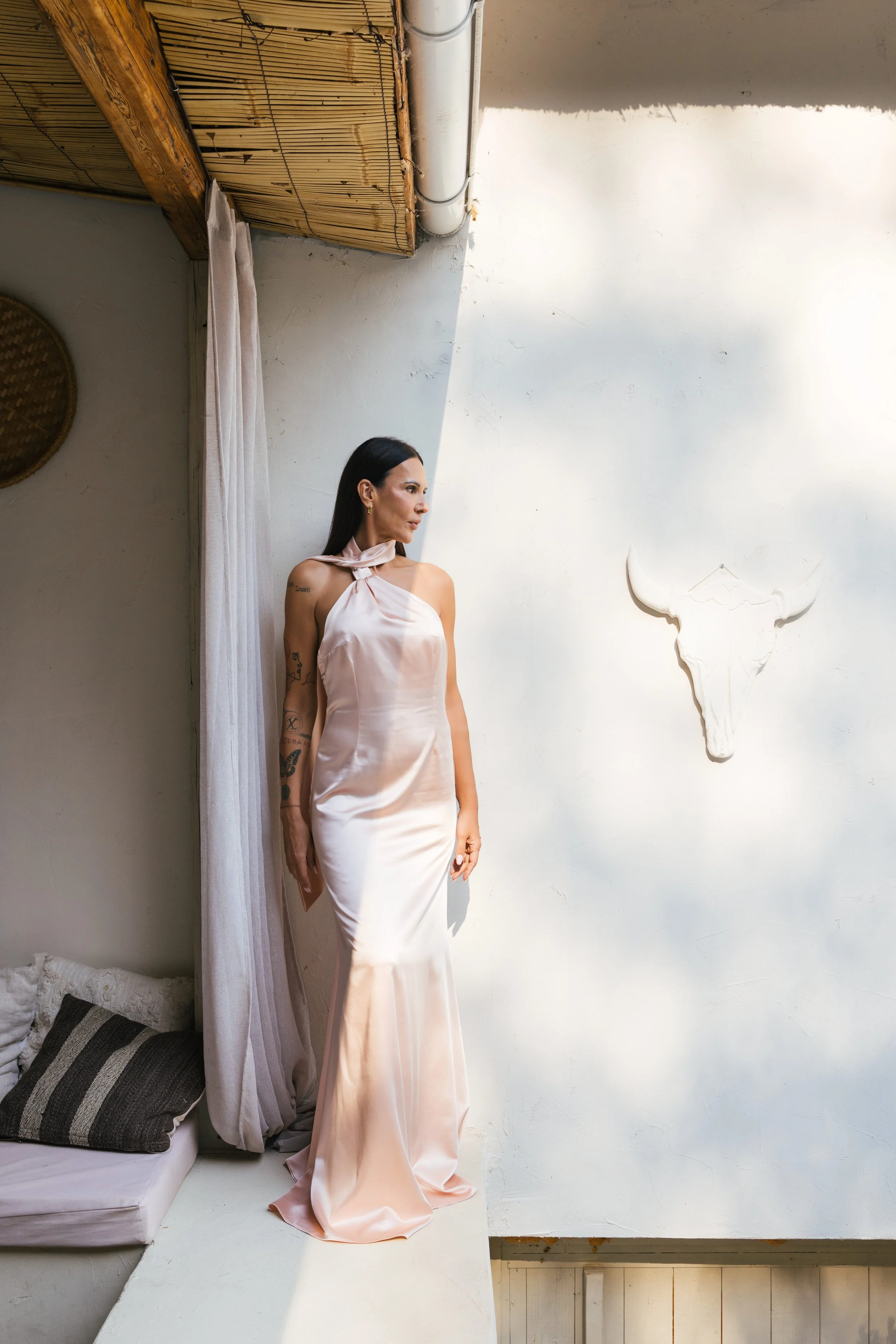 Žena u svetloružičastoj satenskoj haljini stoji pored belog zida sa dekoracijom u obliku lobanje bika, u prostoriji sa pletenim panelima na plafonu i sofom sa prugastim jastucima. Bobana Simovic