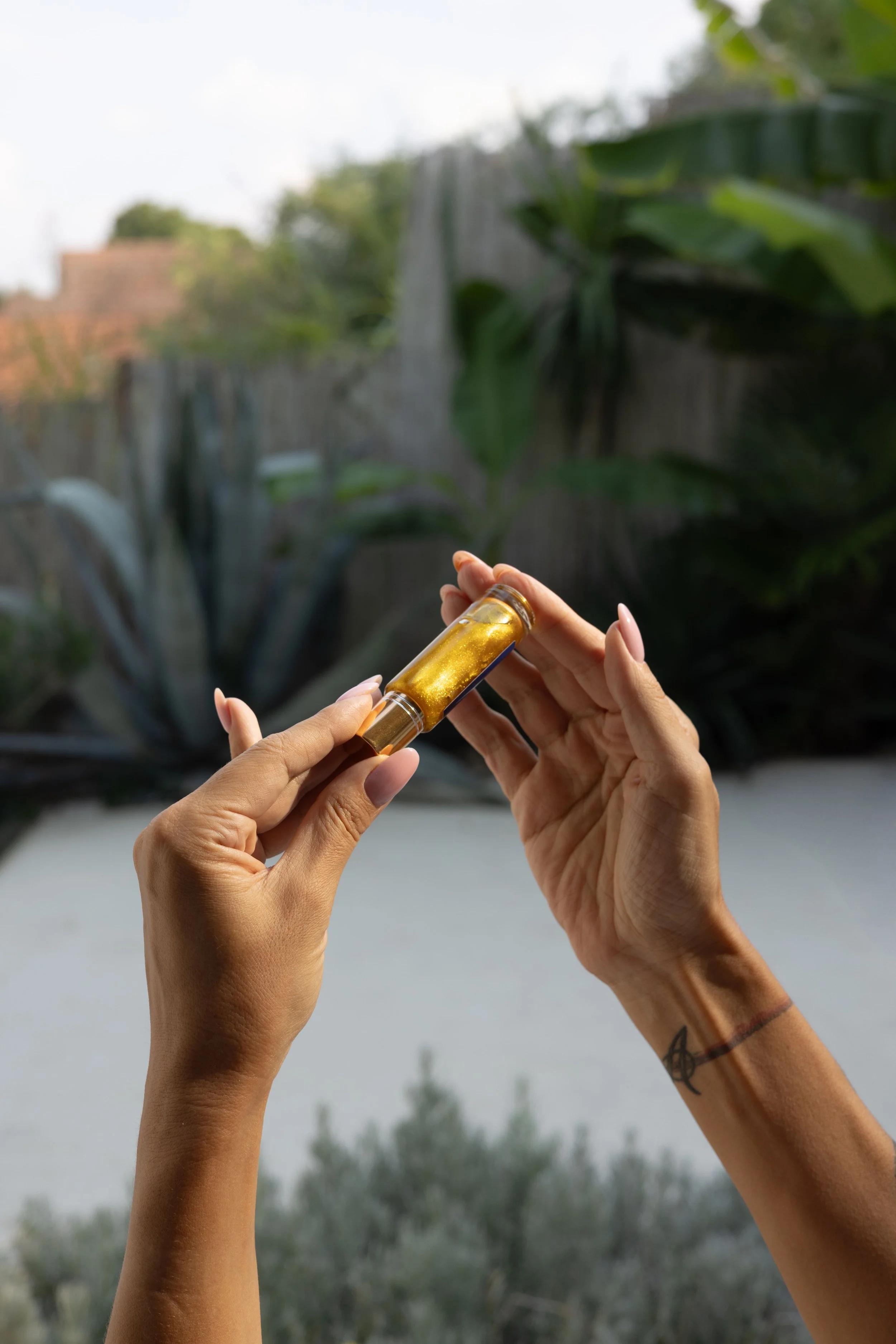 Ruke koje drže malu staklenu bočicu sa zlatnim uljem, ispred prirodnog ambijenta sa krupnim zelenim listovima i vodopadom — susret prirodne frekvencije i ženskog srca.