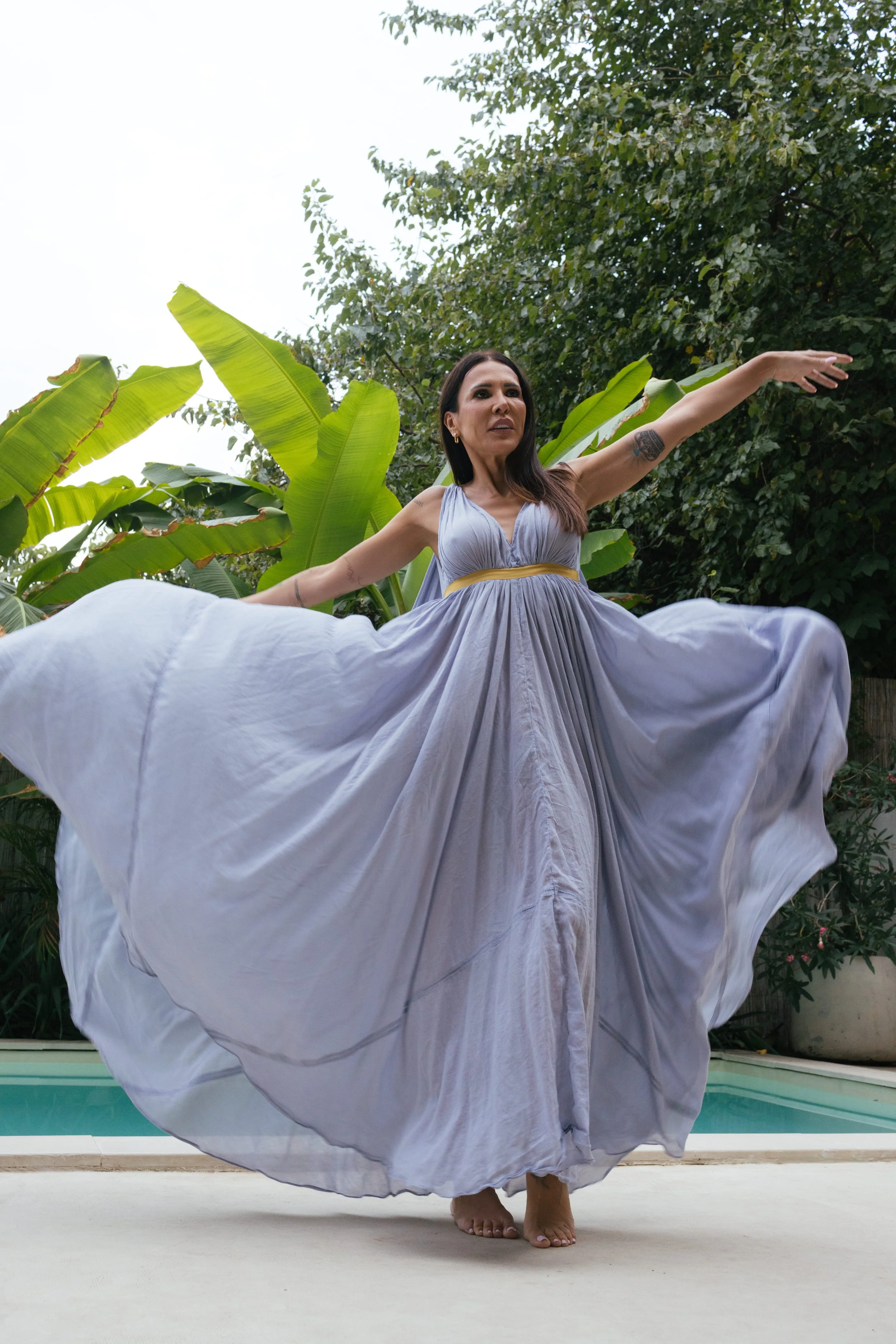 Žena u lepršavoj ljubičastoj haljini sa zlatnim pojasom pleše pored bazena na otvorenom, okružena velikim tropskim lišćem i drvećem. Bobana Simovic