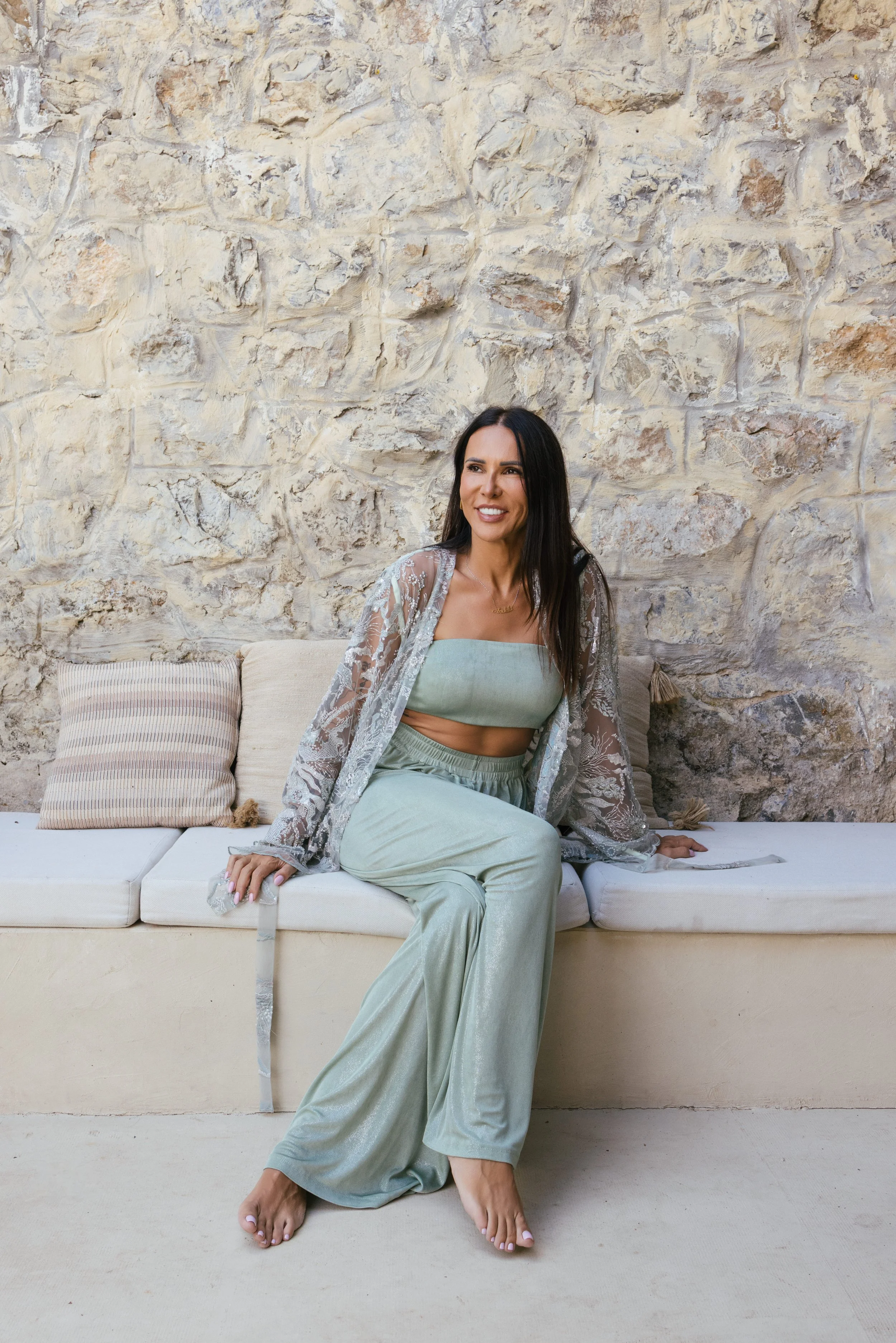 Žena u mint zelenoj odeći za opuštanje, sa prozirnim čipkastim kardiganom, sedi na klupi uz kameni zid, osmehuje se i gleda u stranu. Bobana Simovic