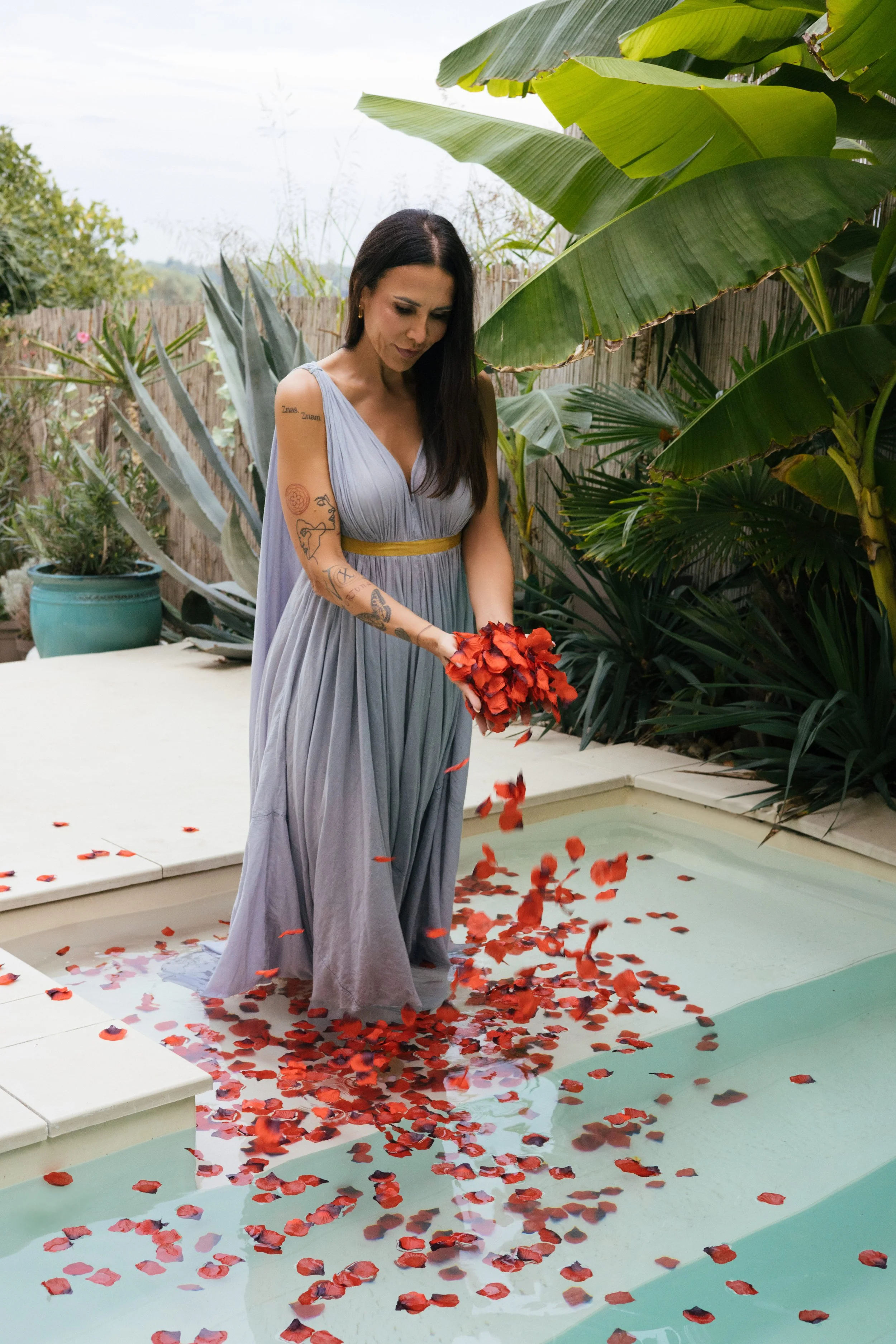 Žena u lepršavoj sivoj haljini sa žutim pojasom stoji u bazenu okružena velikim tropskim lišćem i drži crvene cvetne latice iznad vode. Bobana Simovic