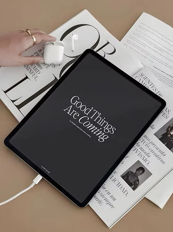 Tablet displaying 'Good Things Are Coming' on a desk with newspapers, a hand holding AirPods, and a sheet of paper.