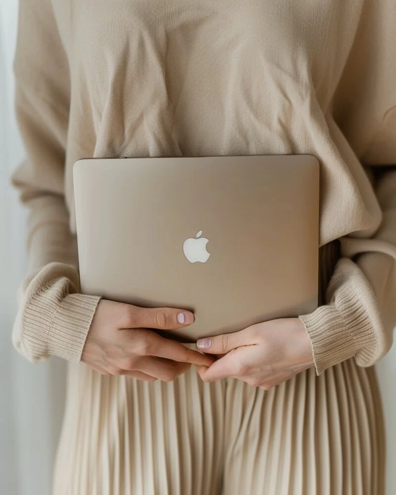 Person holding a closed silver MacBook with Apple logo, dressed in beige sweater and pleated skirt.