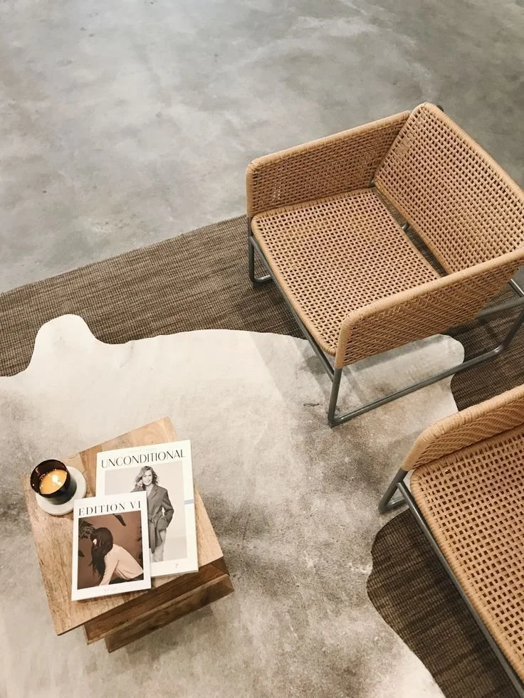 A cozy seating area with two rattan chairs, a wooden side table with magazines, and a small candle on a white plate, all on a concrete floor.