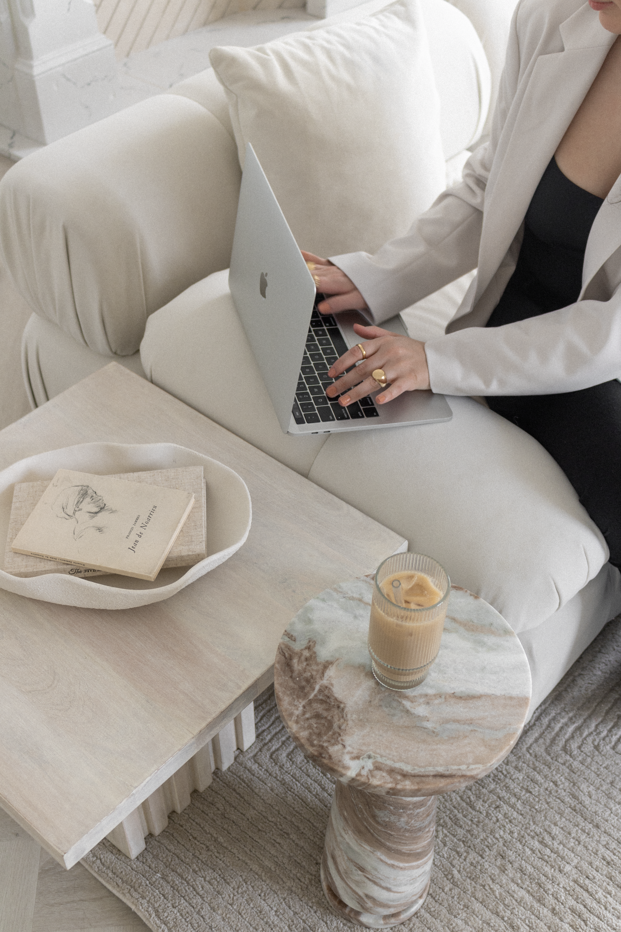 Person sitting on a white sofa using a laptop, with a book in a white bowl, a glass of iced coffee on a marble side table, and a beige rug underneath.