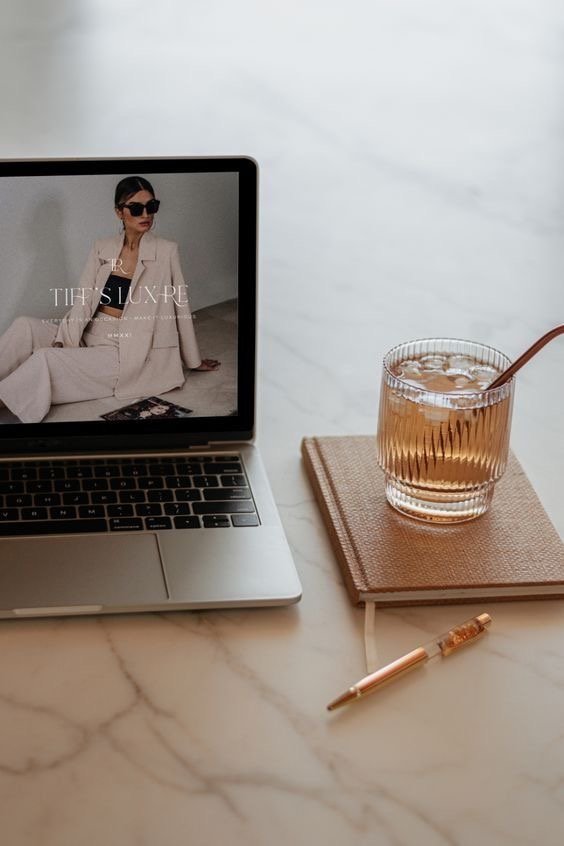 A flat lay of a workspace featuring a laptop displaying a woman in a suit and sunglasses, a glass of iced tea with a straw on a pink book, a closed notebook, and a gold pen on a marble surface.
