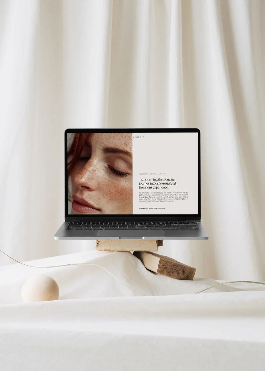 A laptop placed on a stack of round and rectangular stone blocks, displaying a webpage with a close-up of a woman's face with freckles and closed eyes on the screen. The background features a cream-colored curtain and a minimalist, beige decorative sphere and orb on a white fabric surface.