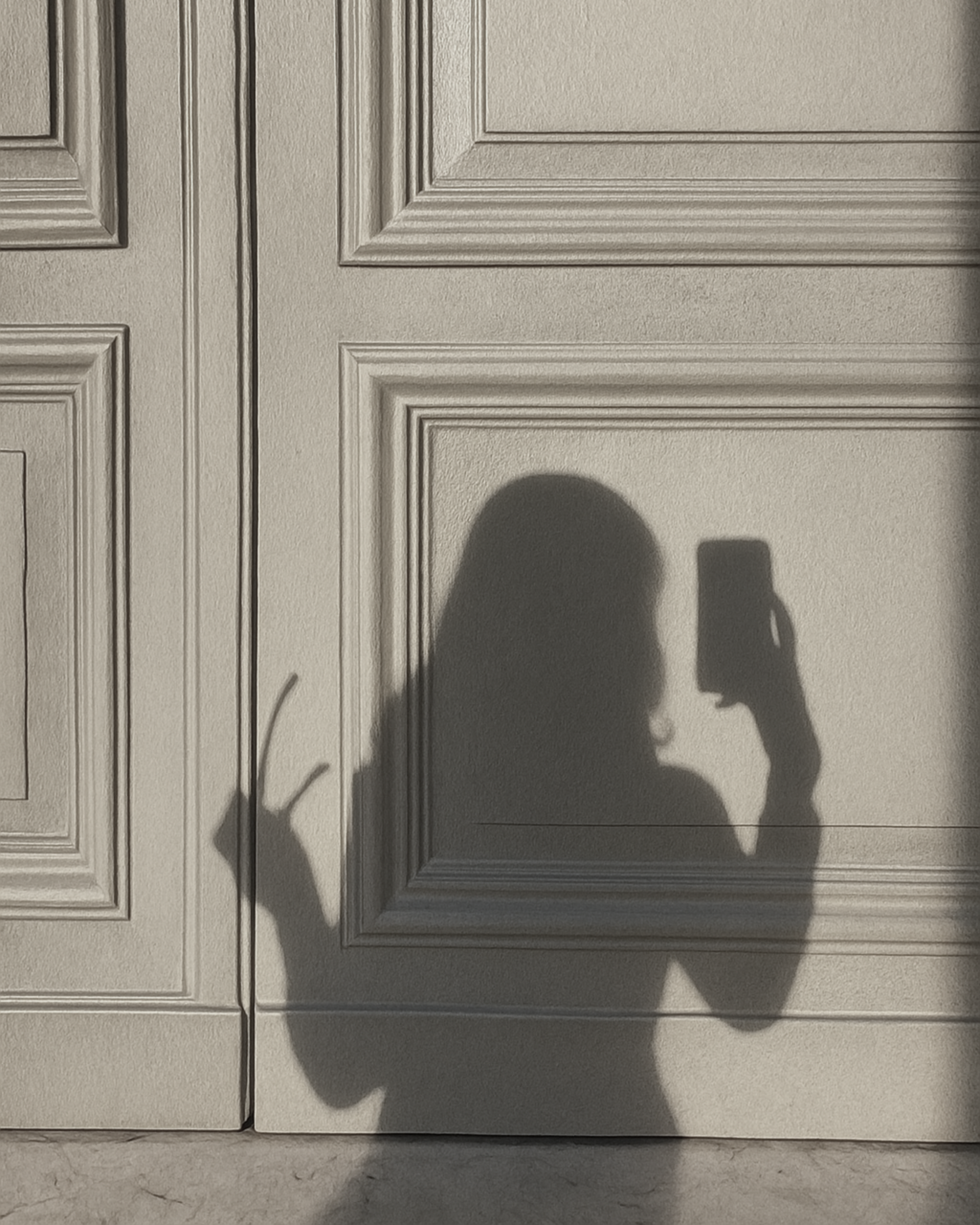 Shadow of a woman holding a phone to take a picture, with one hand making a peace sign, projected on a wall with decorative wood paneling.