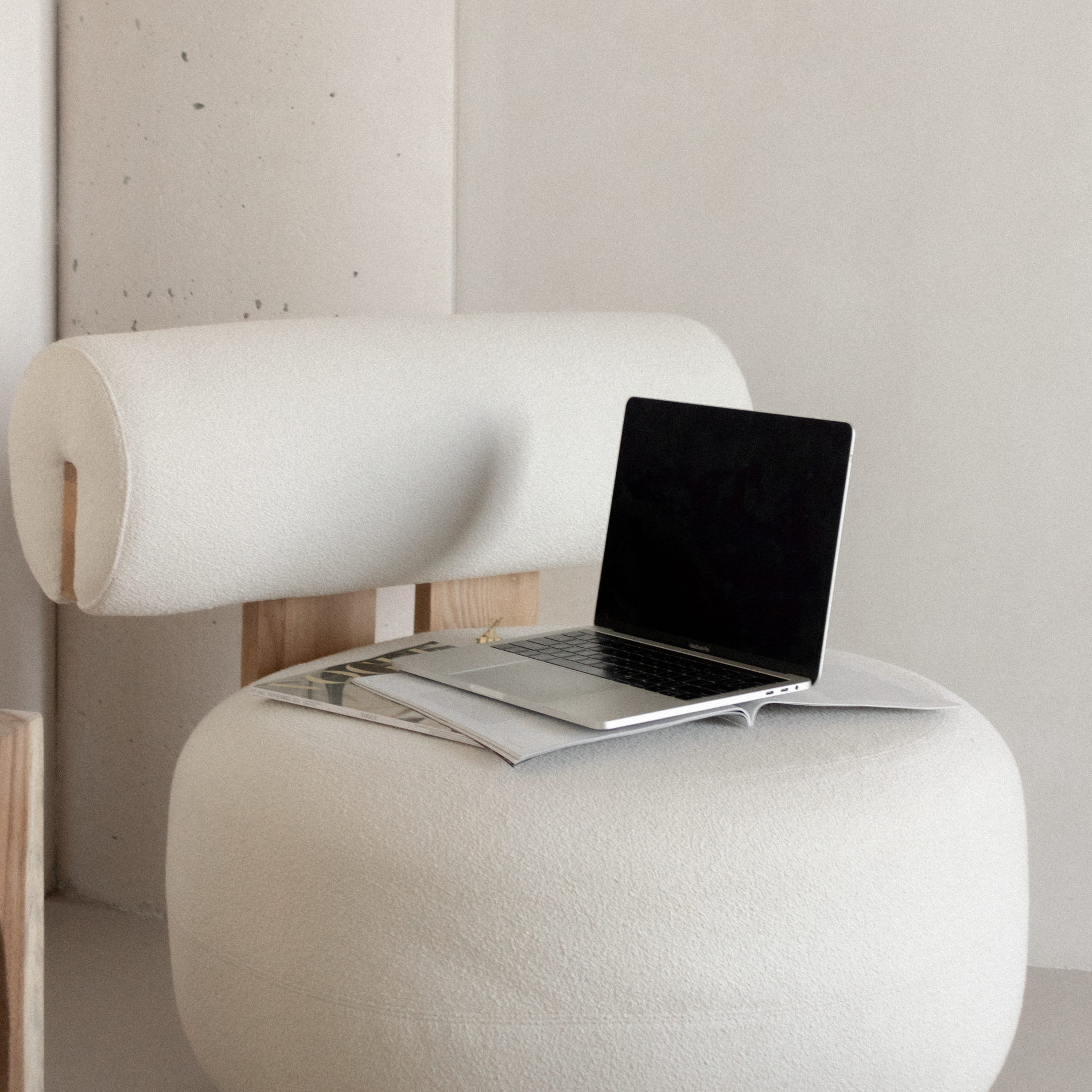 A white modern chair with a rolled backrest, on which a silver MacBook laptop and an open magazine are placed.