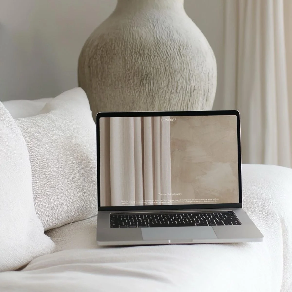 A laptop sits on a white sofa with textured cushions. Behind it, there is a large textured beige vase and a cream-colored curtain.