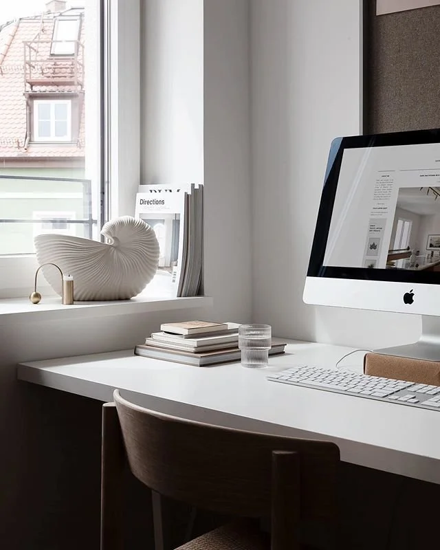 A minimalist workspace featuring a white desk, a computer monitor, a glass of water, and a stack of books. On the windowsill, there is a decorative seashell sculpture, some magazines or manuals, and a directions booklet. The room has natural light coming through a window, revealing a building facade outside.