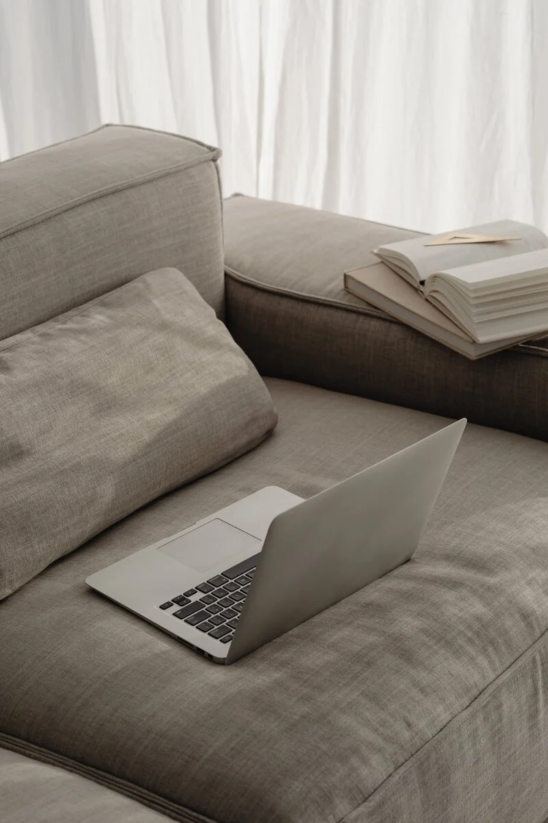 A gray sofa with a laptop open on it, next to a stack of open books and papers, in front of white curtains.