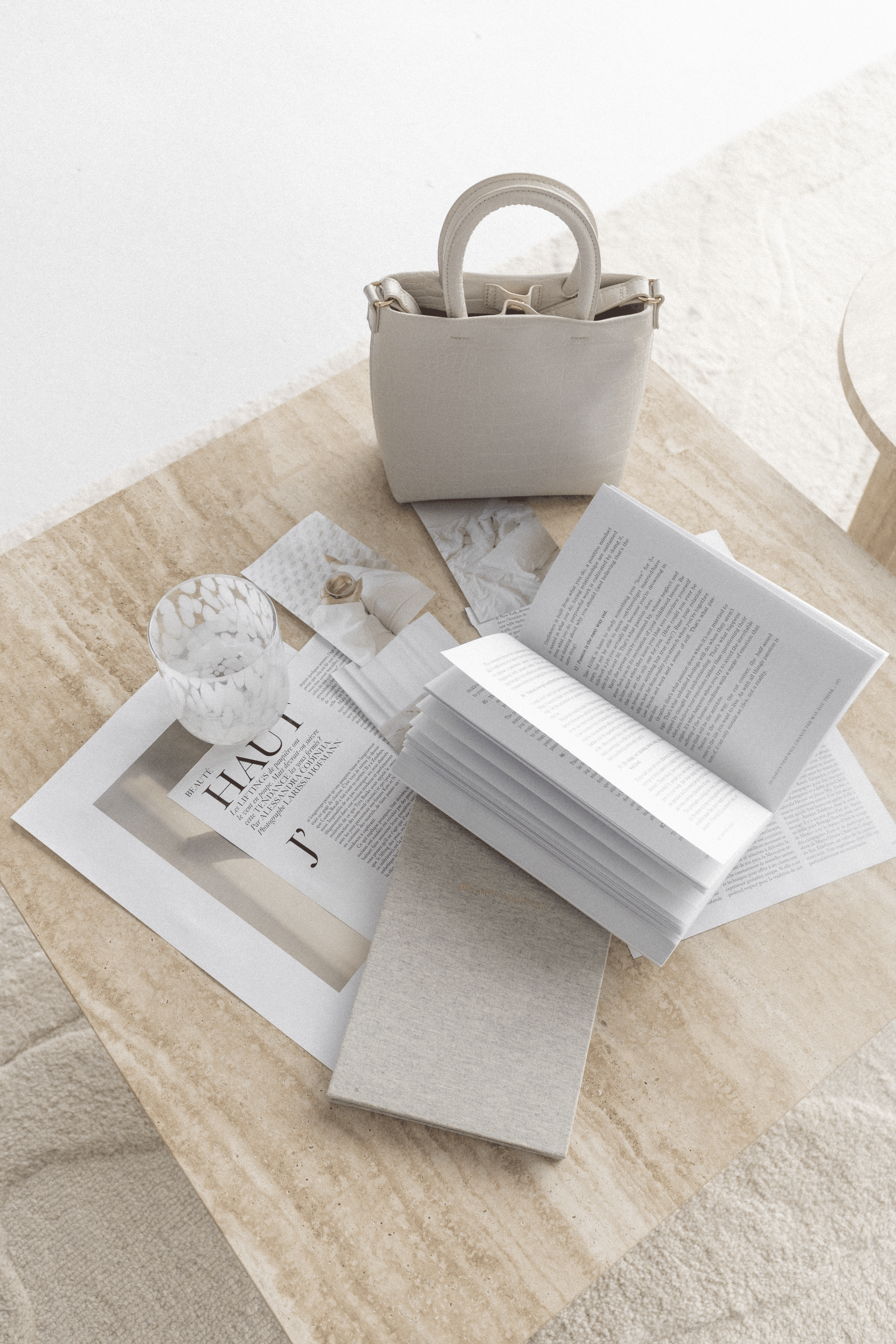 A beige handbag on a wooden table surrounded by open books, papers, a glass with a decorative pattern, and a fabric-covered notebook.