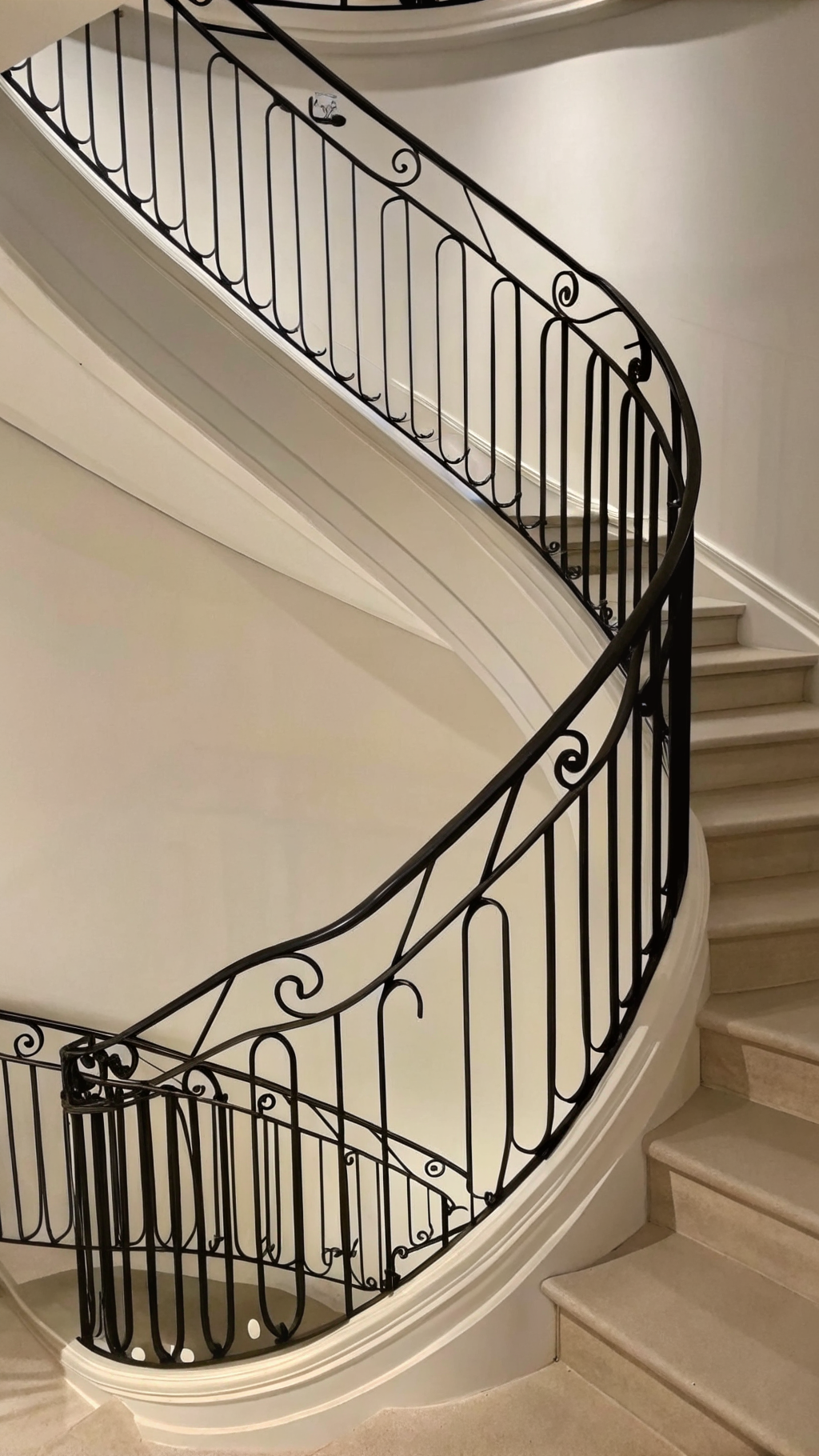 Photo of a curved staircase with beige marble steps and an intricate black wrought iron railing.