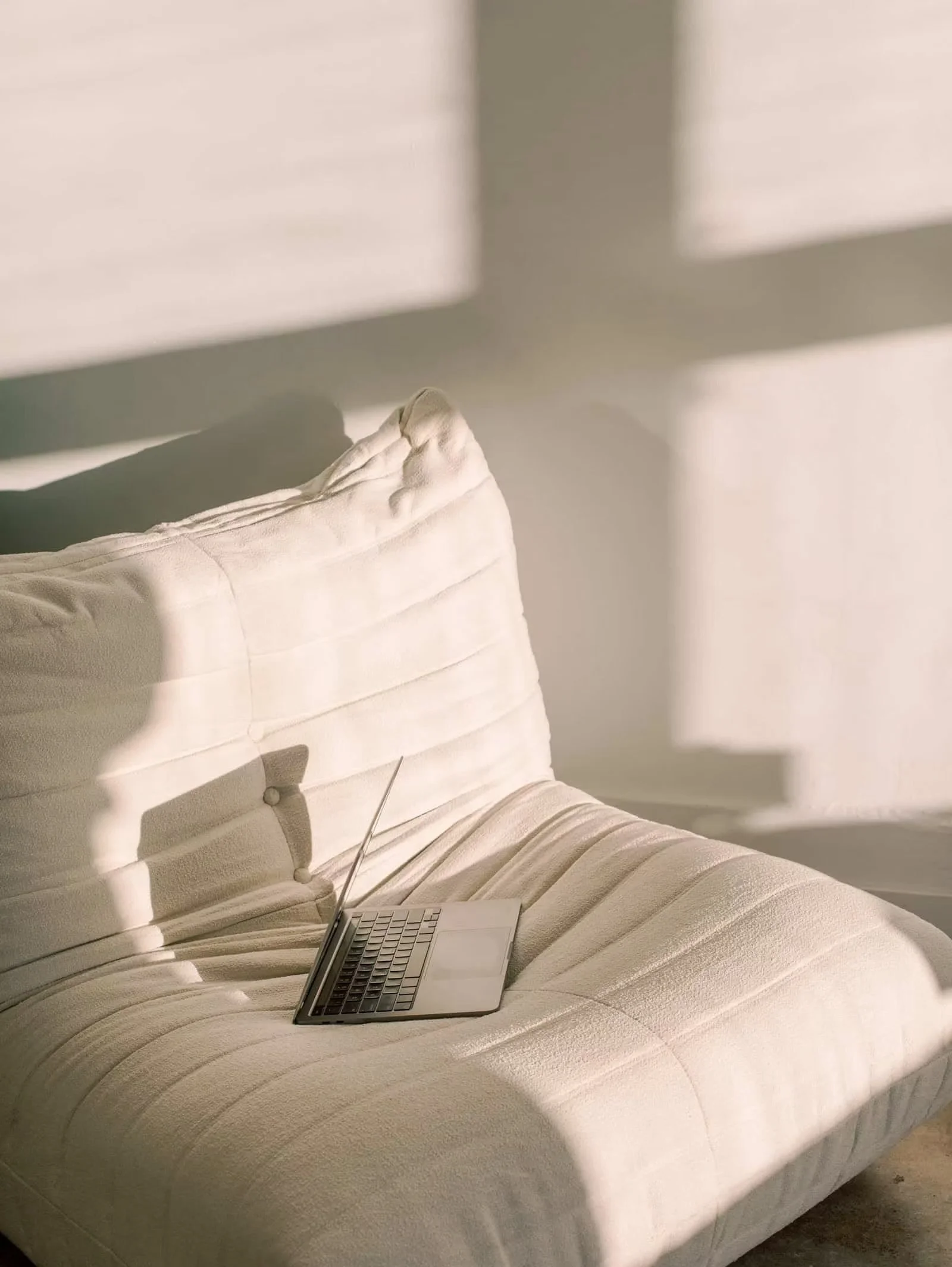 Sunlight casts shadows on a cream-colored sofa with a small silver laptop resting on it.