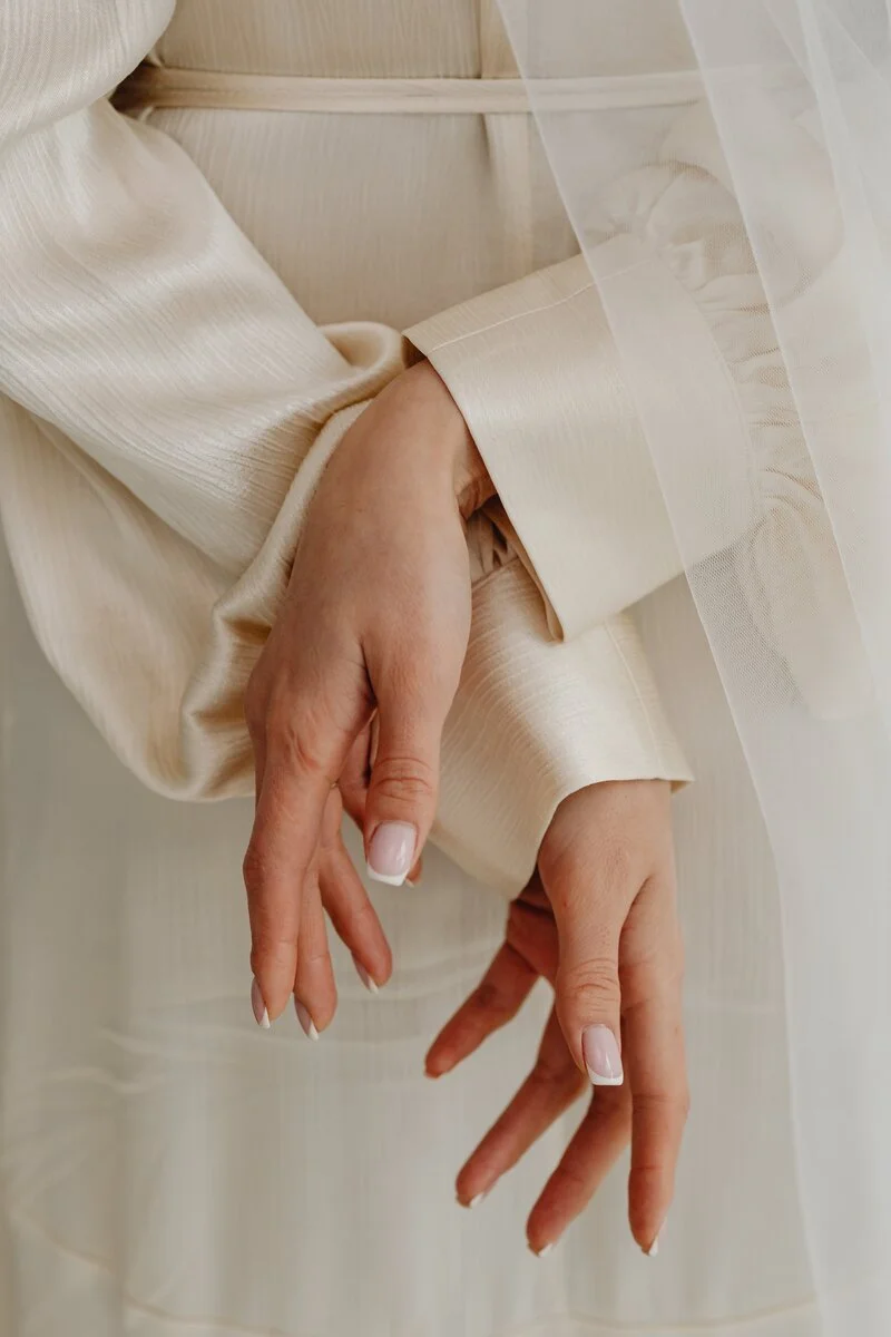 Close-up of a person wearing elegant, cream-colored satin or silk long-sleeve clothing, with hands gracefully posed and hanging down, partially covered by a sheer, white fabric or veil.