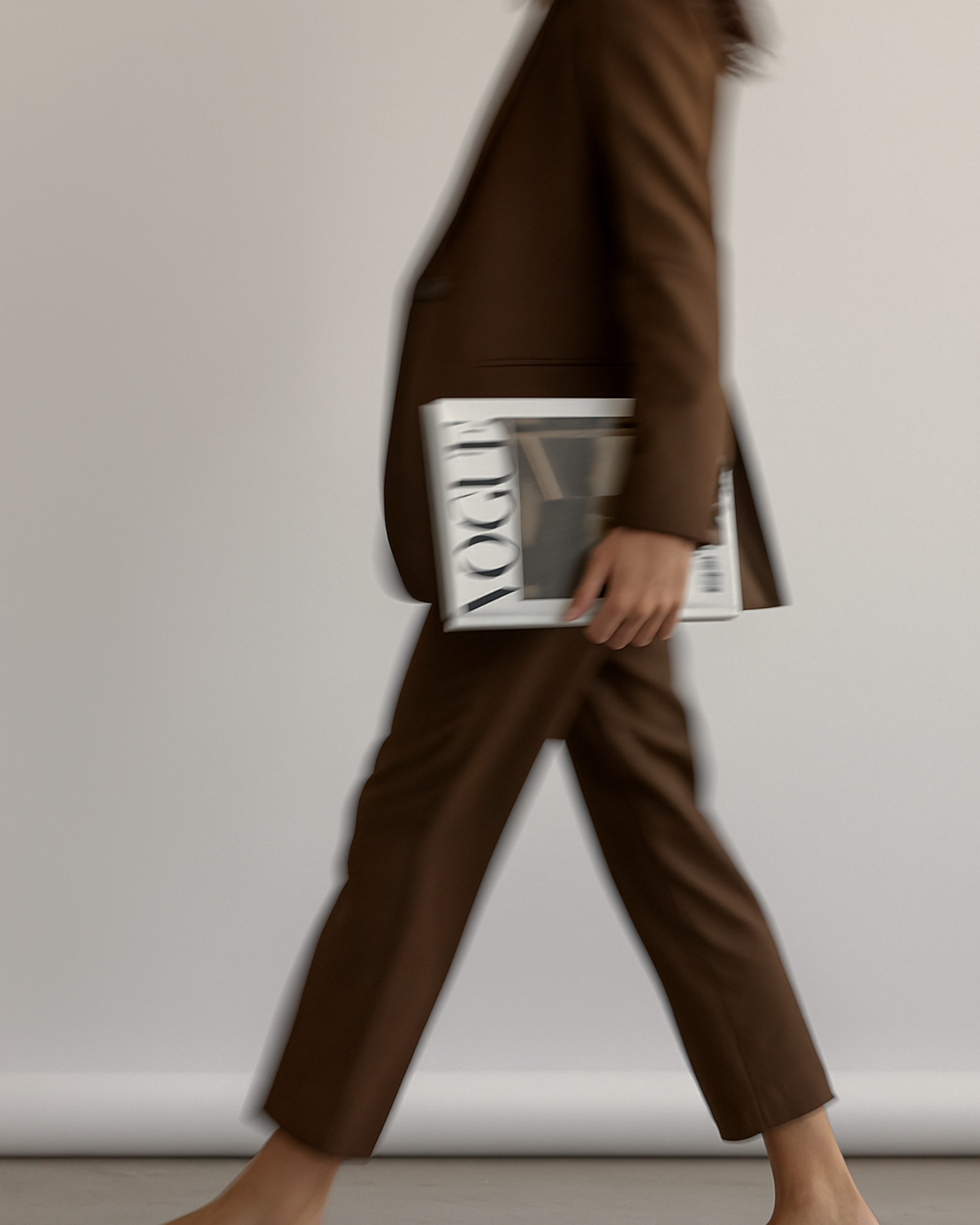 A person walking in a brown suit, holding a magazine or catalog called 'YOOX' in their right hand, with a plain gray background.