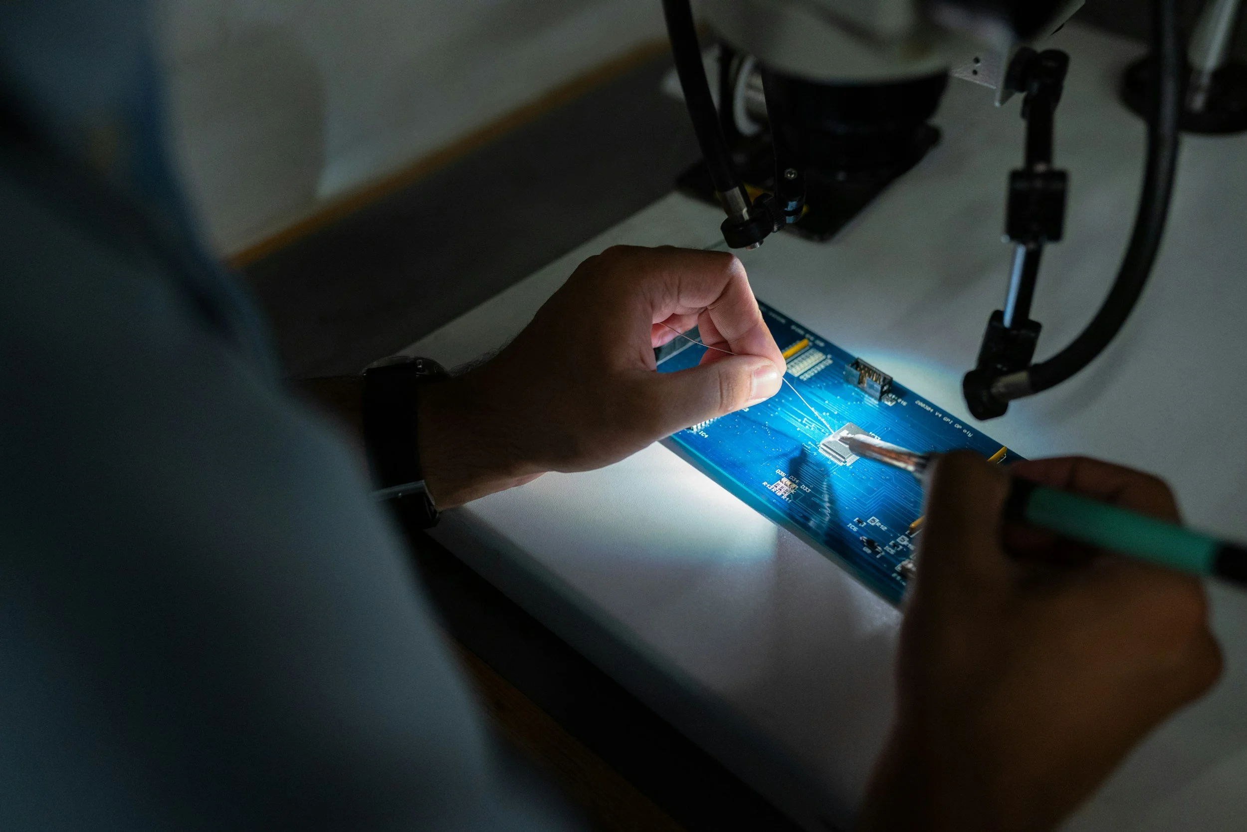 Person working on a blue circuit board using a soldering iron, under a microscope in a lab setting.