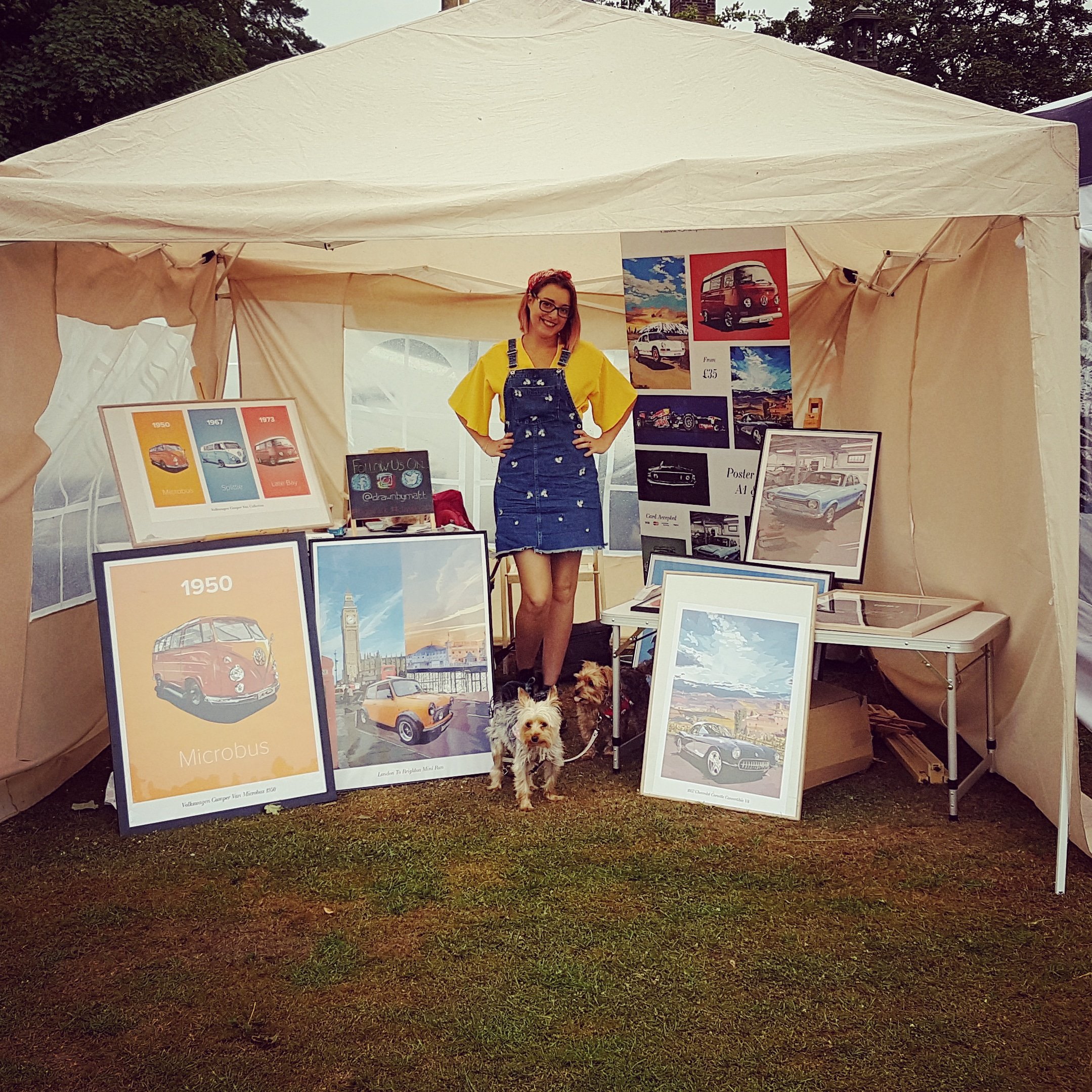 An outdoor event with a beige canopy tent displaying vintage car posters, artwork, and a smiling woman with red hair, glasses, yellow shirt, and denim overalls, standing in front with two small dogs at her feet, on a grassy area with trees in the background.