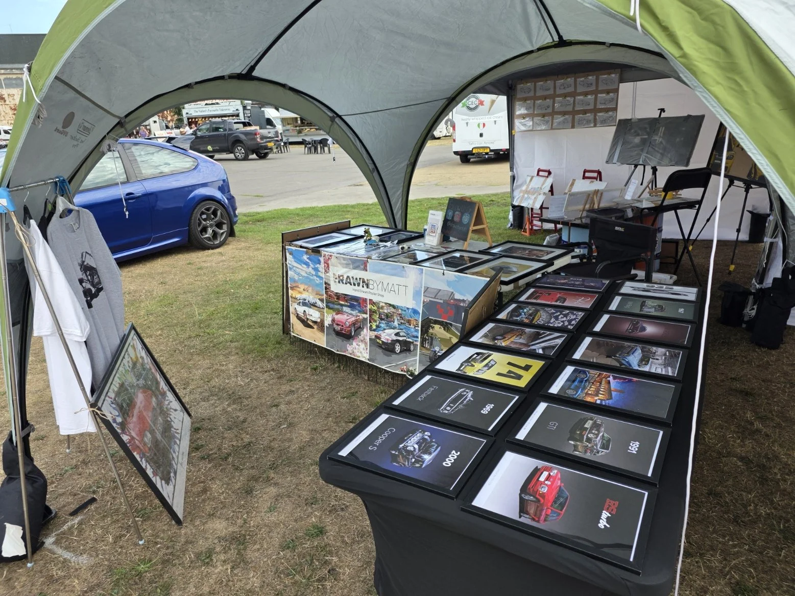 An outdoor tent setup displaying framed photographs and art prints of cars, with some framed pictures on a table inside and a blue car parked nearby.