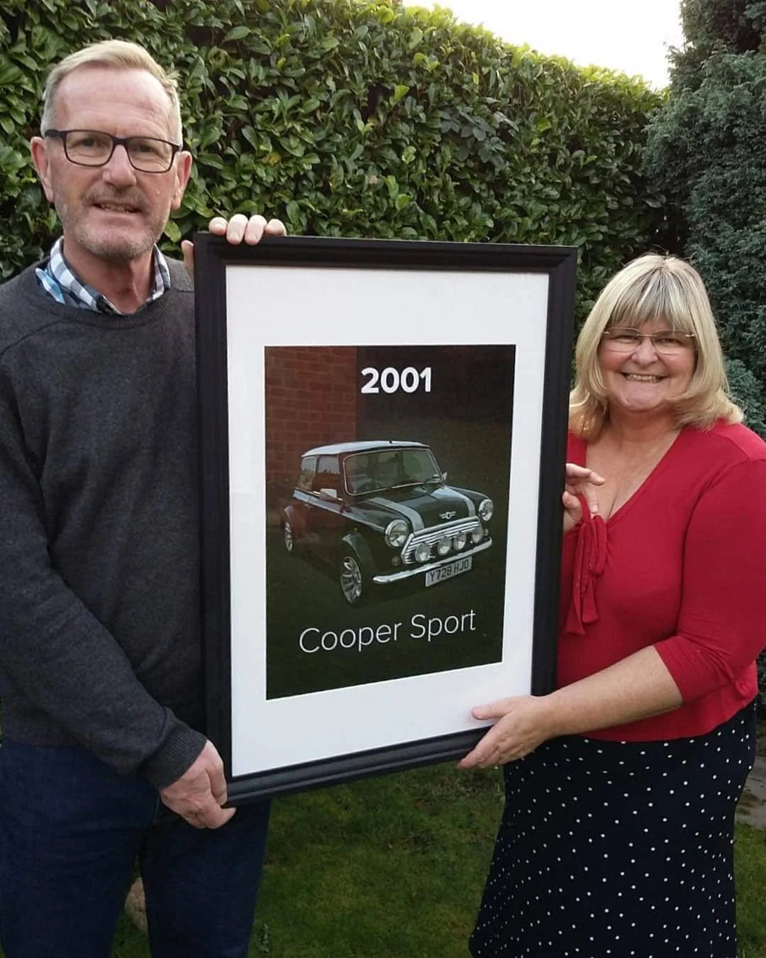 A man and a woman are smiling while holding a framed picture of a vintage Mini Cooper car from 2001, labeled 'Cooper Sport,' outdoors in a garden.
