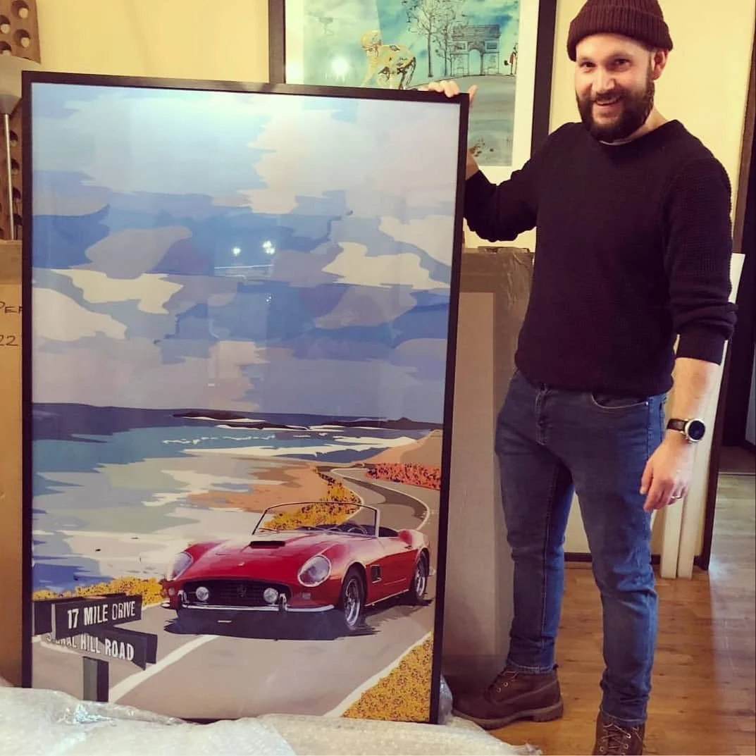 A man wearing a maroon beanie, black sweater, and jeans standing next to framed art depicting a red vintage car on a coastal road with ocean, hills, and cloudy sky background.