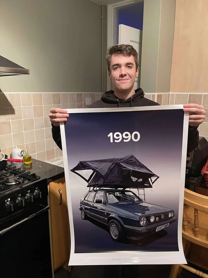 A young man smiling while holding a poster showing a 1990 car with a roof tent and an umbrella attached, inside a kitchen.